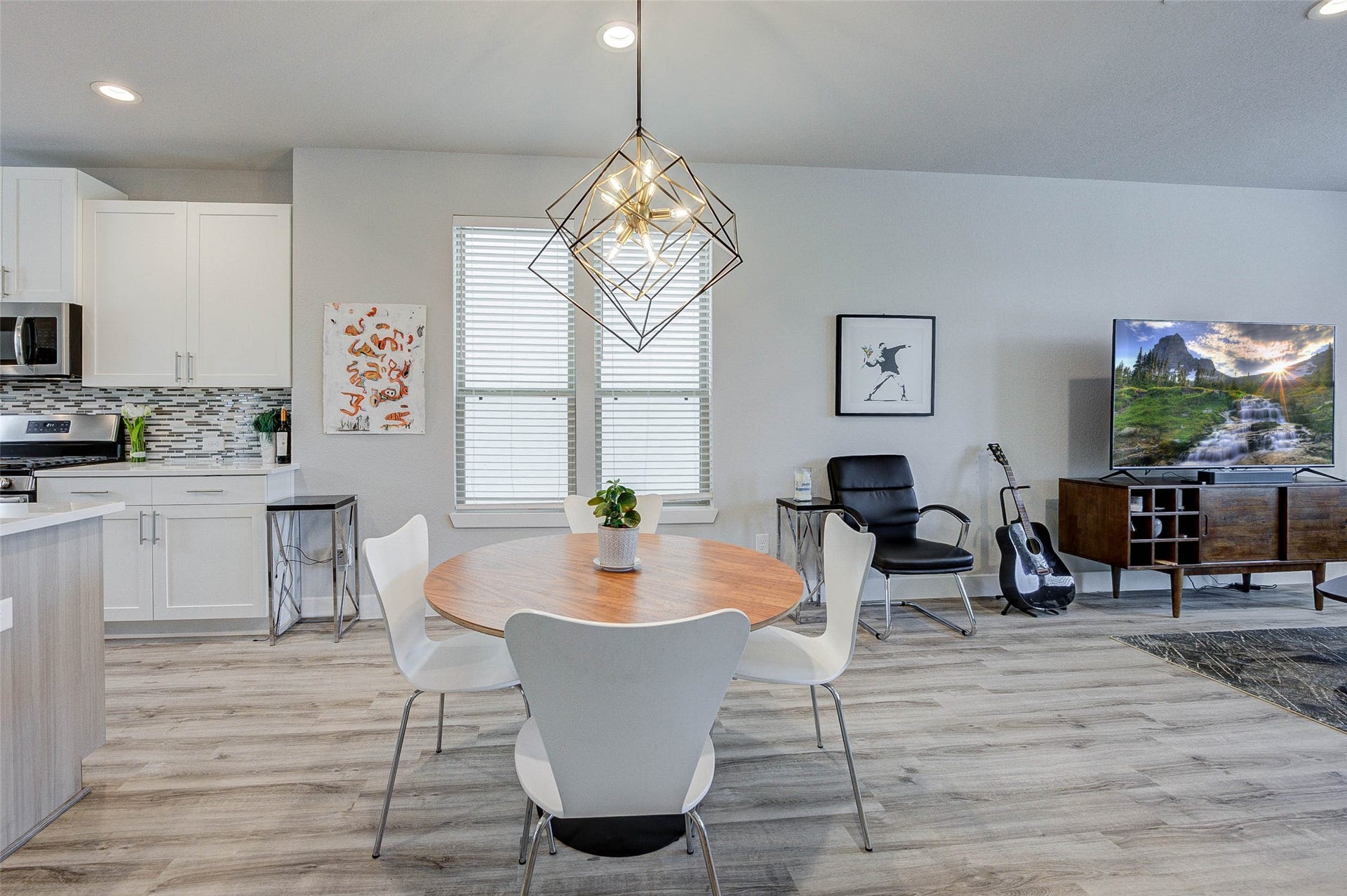 18732 Centro Main Street, Shenandoah, TX 77385 This modern open-plan living area features a sleek kitchen with stainless steel appliances and white cabinetry. It seamlessly flows into a cozy dining space with a stylish geometric light fixture. The living area includes a comfortable chair, TV setup, and contemporary decor, all set on elegant wood flooring.