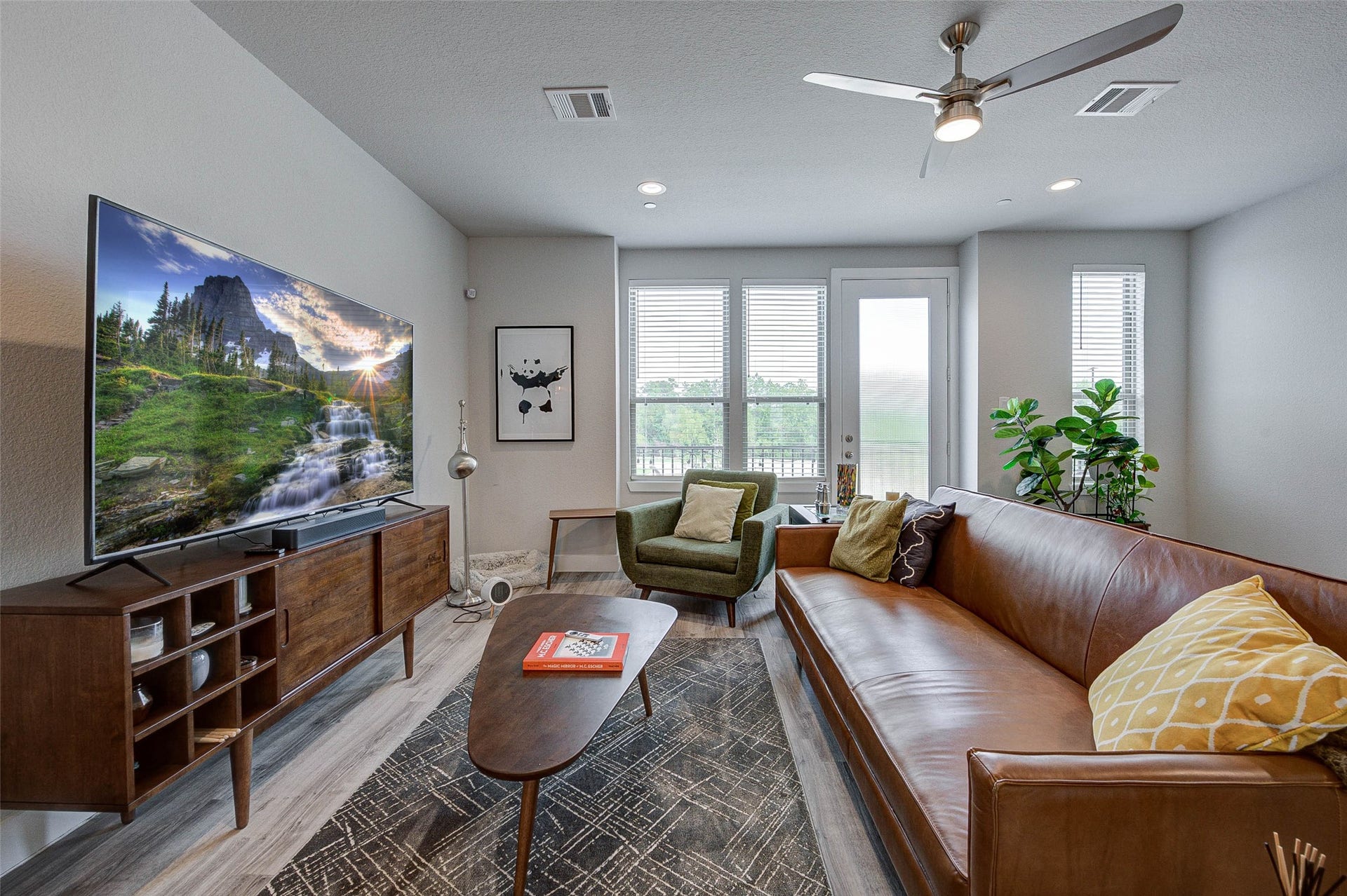 18732 Centro Main Street, Shenandoah, TX 77385 This cozy living room features a modern design with a sleek ceiling fan, a large TV, and a stylish wooden media console. The space is brightened by natural light from large windows and a door leading to an outdoor area. Comfortable seating includes a brown leather sofa and an olive armchair, complemented by a patterned rug and decorative pillows.