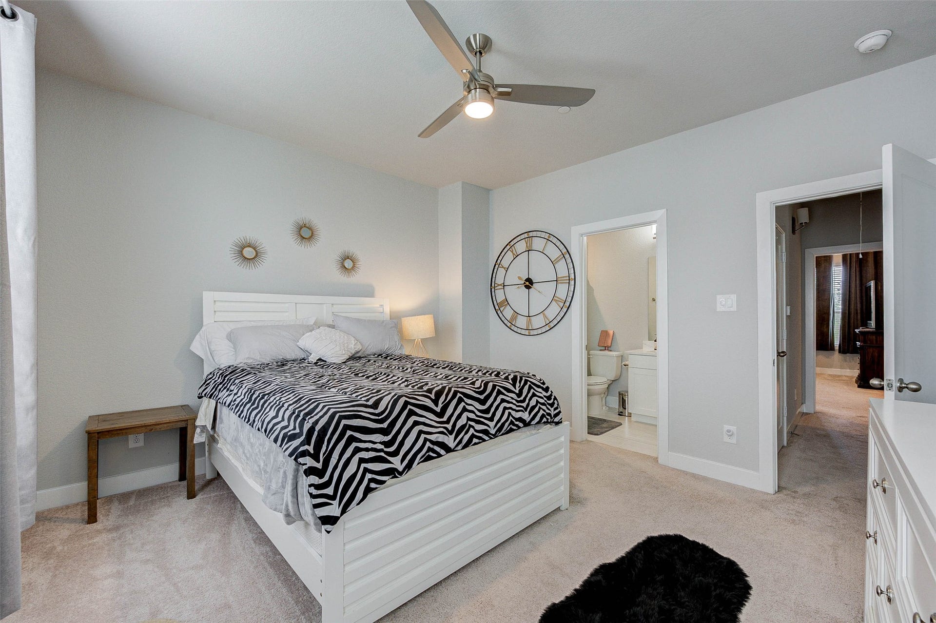 18732 Centro Main Street, Shenandoah, TX 77385 This is a cozy and bright bedroom with a modern ceiling fan, neutral walls, and carpeted flooring. It features a large wall clock, stylish decor, and an en-suite bathroom, providing both functionality and style.