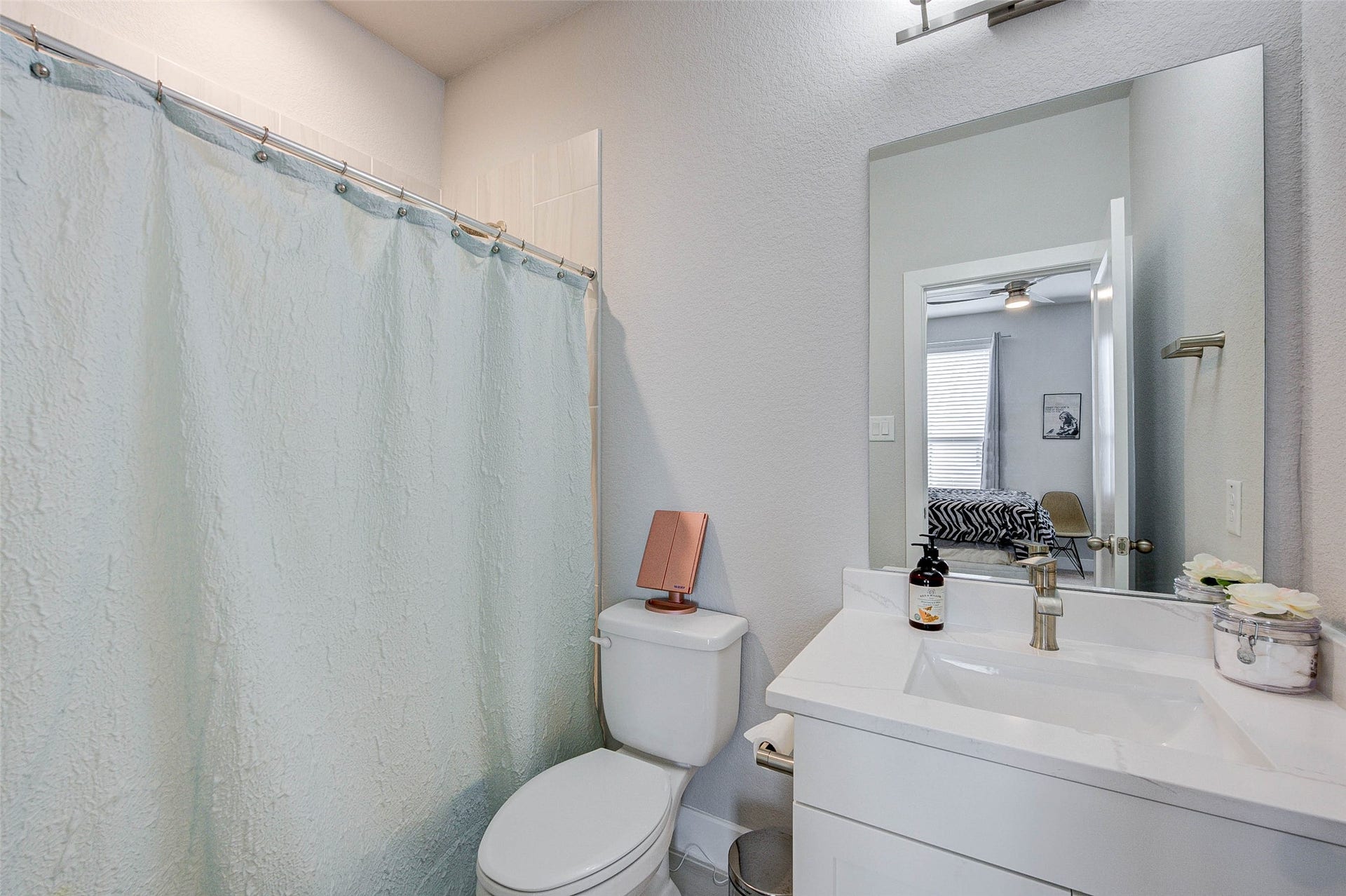 18732 Centro Main Street, Shenandoah, TX 77385 This bathroom features a clean, modern design with a white vanity and sink, complemented by a large mirror. It includes a toilet and a shower with a light-colored curtain. The room is well-lit, enhancing its fresh and airy feel.
