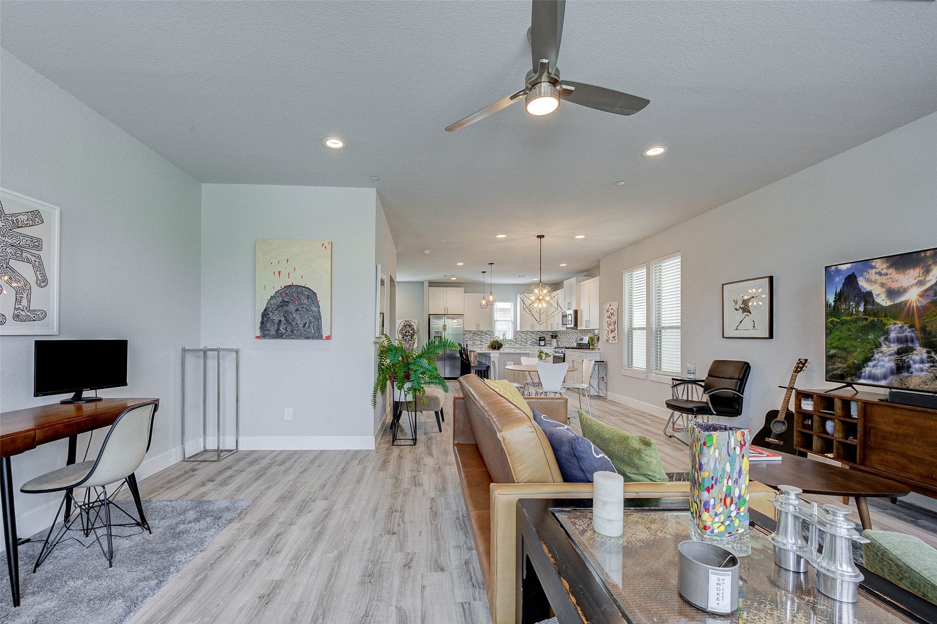 18732 Centro Main Street, Shenandoah, TX 77385 This modern open-concept living space features a cozy seating area with a stylish ceiling fan, a work desk, and a large TV. The kitchen is in the background with sleek cabinetry, stainless steel appliances, and a central island. The room is well-lit with natural light from multiple windows, and there are tasteful decorative touches throughout.