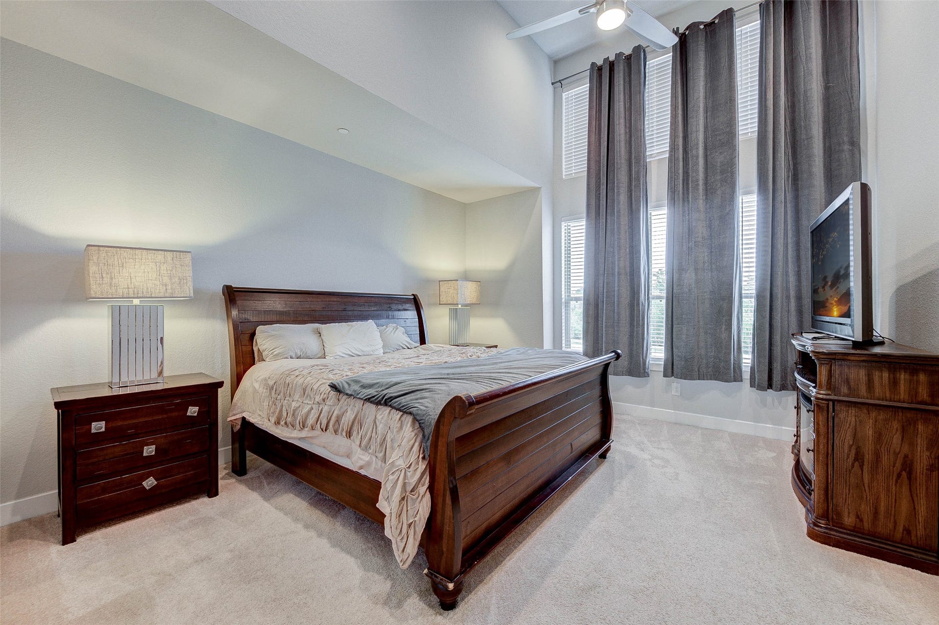 18732 Centro Main Street, Shenandoah, TX 77385 This spacious bedroom features a high ceiling, large windows with elegant drapes, and abundant natural light. It includes a wooden bed set with matching nightstands and a dresser, creating a warm and inviting atmosphere.