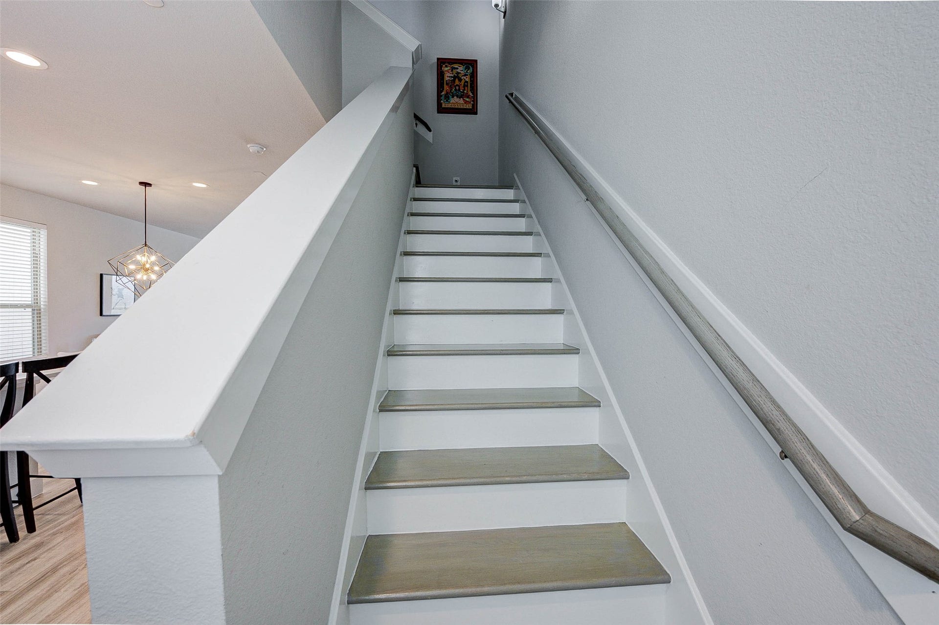 18732 Centro Main Street, Shenandoah, TX 77385 This photo shows a modern staircase with light gray steps and a sleek handrail. The walls are painted a soft gray, and there's a glimpse of a dining area with contemporary lighting. A small piece of artwork hangs at the top of the stairs.