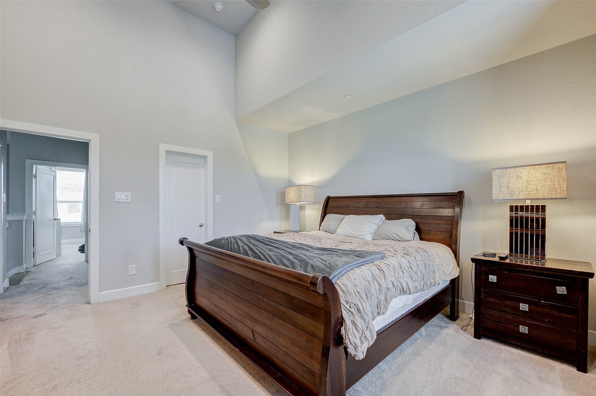 18732 Centro Main Street, Shenandoah, TX 77385 This spacious bedroom features a high ceiling and soft carpeting, creating an inviting atmosphere. It includes a large wooden bed with matching nightstands and modern lamps, offering a comfortable and stylish retreat. A doorway leads to additional living spaces, enhancing the room's functionality.