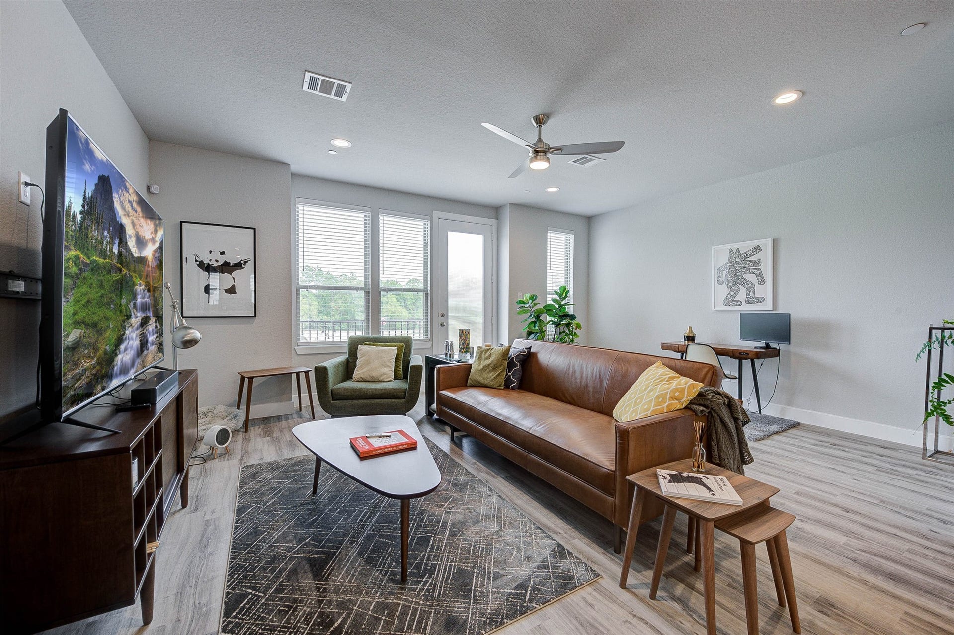 18732 Centro Main Street, Shenandoah, TX 77385 Bright and modern living room with large windows, featuring a stylish leather sofa, sleek TV setup, and a small work desk. The space is accented with plants and contemporary artwork, offering a cozy yet sophisticated atmosphere.