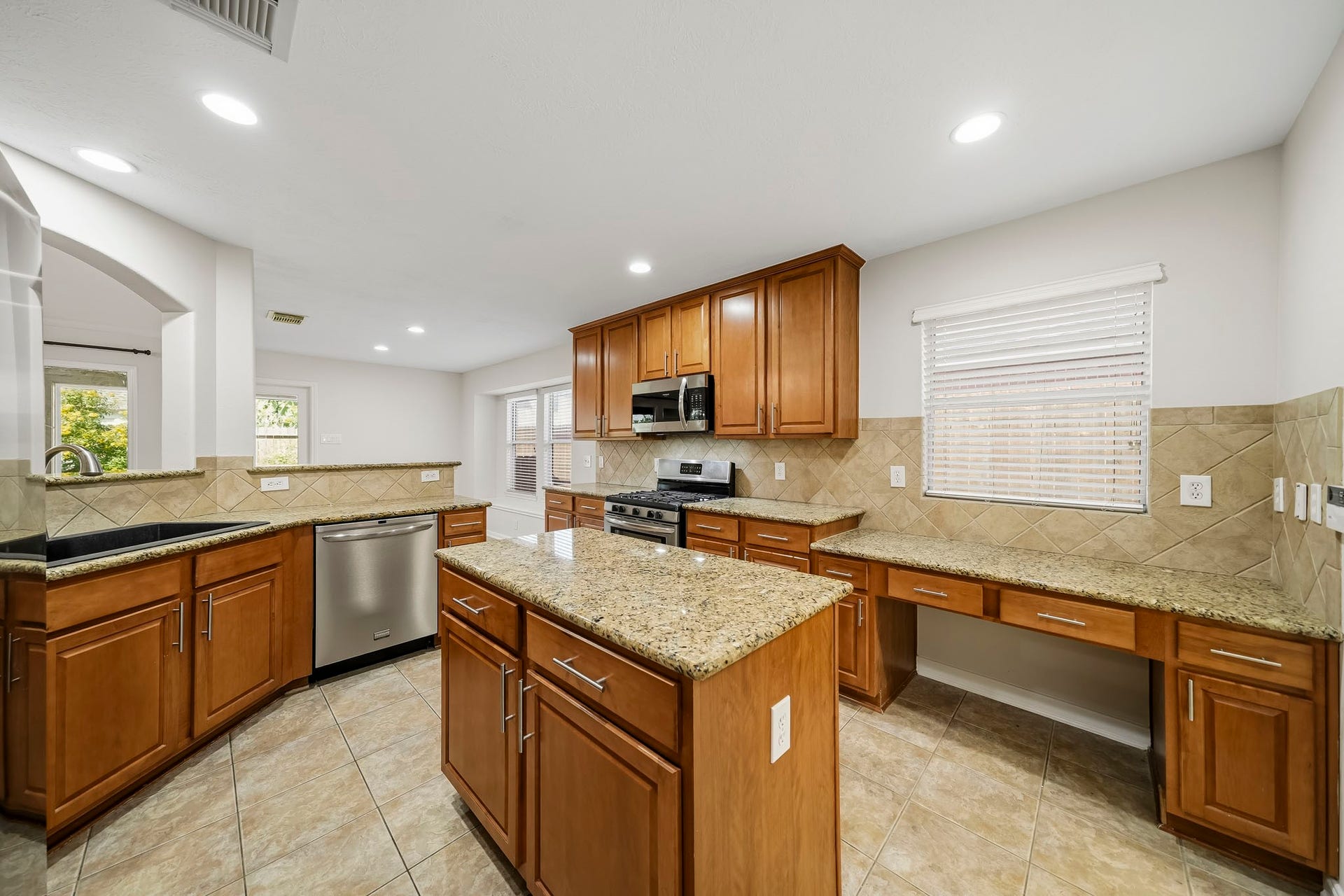 Kitchen features a desk area with a large window, tile backsplash, modern finishes, and stainless steel appliances. This bright space is perfect for work-from-home tasks, offering ample natural light and a seamless transition between culinary prep and productivity.