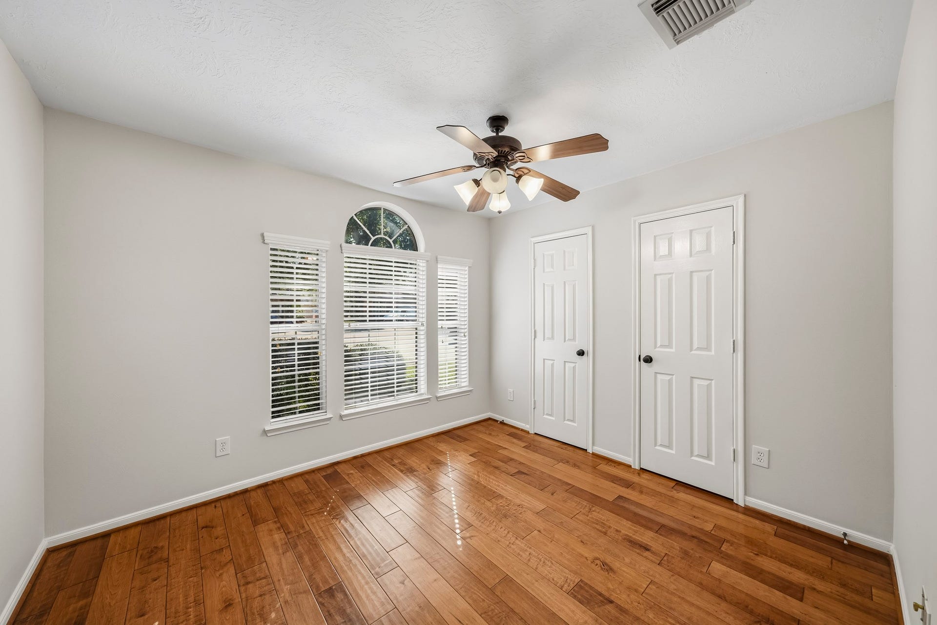 Spacious secondary bedroom featuring gleaming hardwood floors, dual closets, a modern lighted ceiling fan, and elegant windows accentuated by a sophisticated architectural arch.