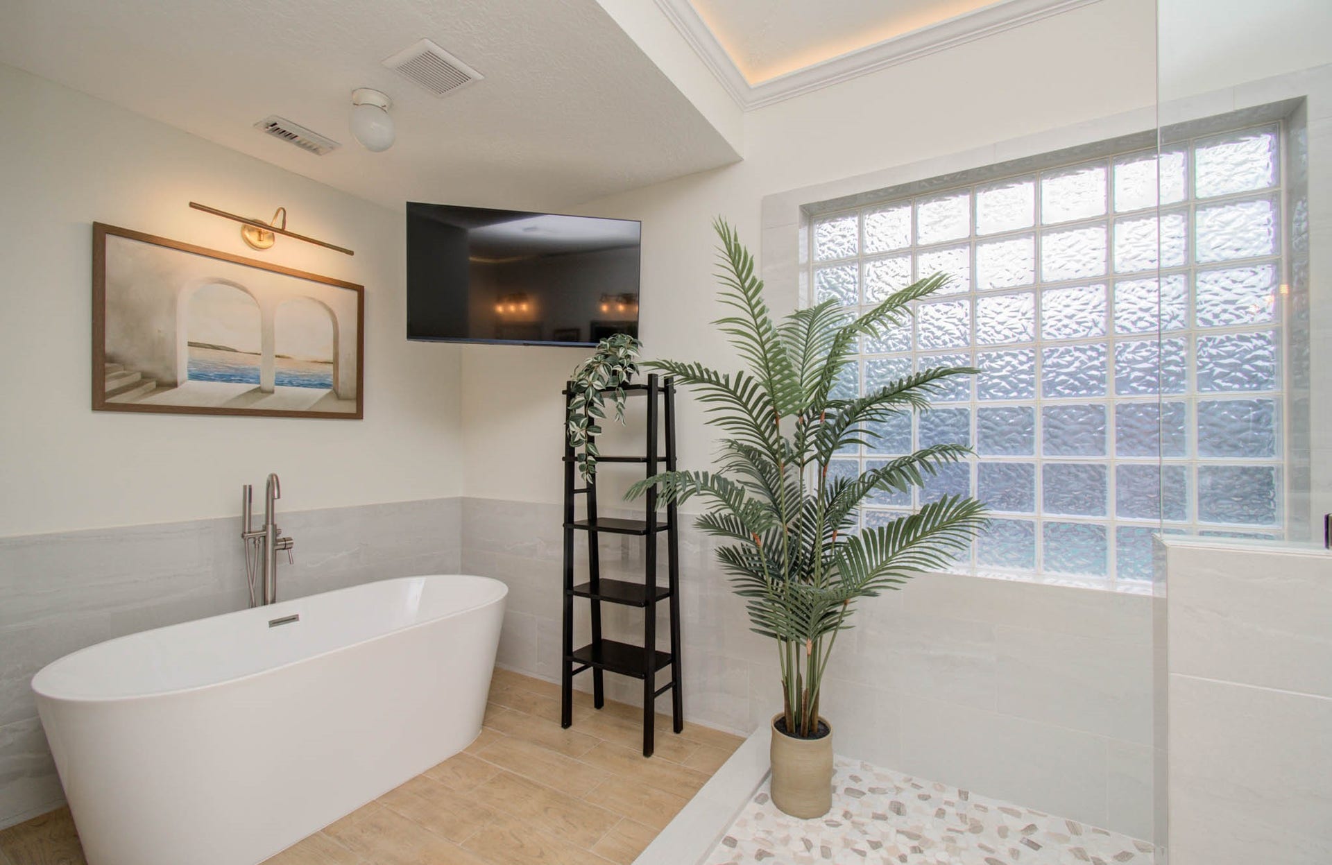 15627 Twisting Springs Drive, Cypress, TX 77433 Luxurious bathroom featuring a freestanding soaking tub, elegant tiled floors, and a large glass block window for natural light. A modern ladder shelf with greenery and a wall-mounted TV add both style and function.