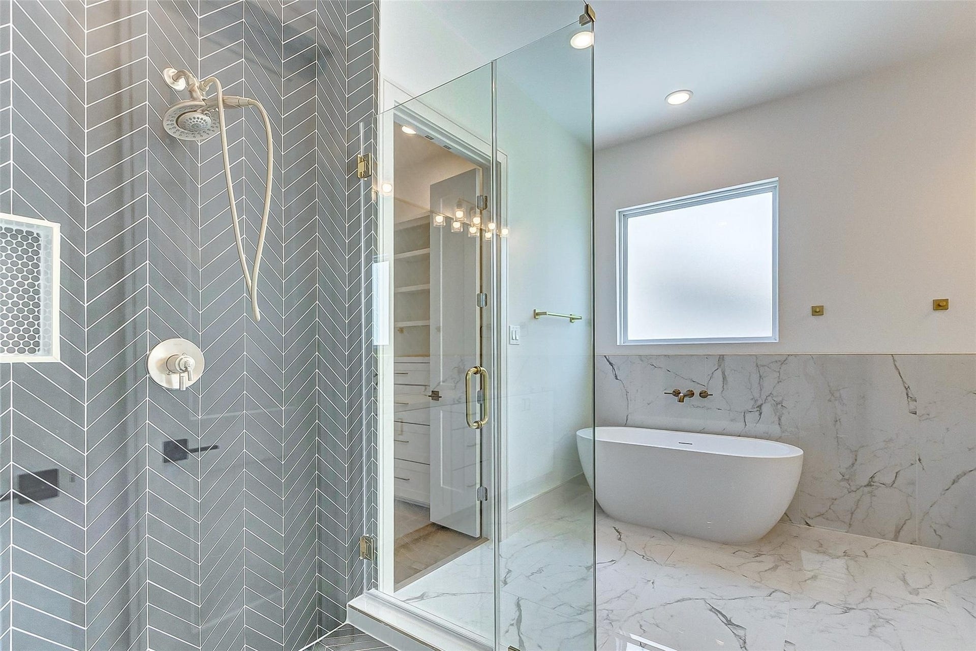 605 E 26th A Street, Houston, TX 77008 Upscale bathroom featuring a luxurious freestanding soaking tub, modern glass-enclosed frameless shower with sleek herringbone tiles, linen closet, dual vanities, large mirror, very spacious walk in closet, private commode, and bright natural light through a frosted window.