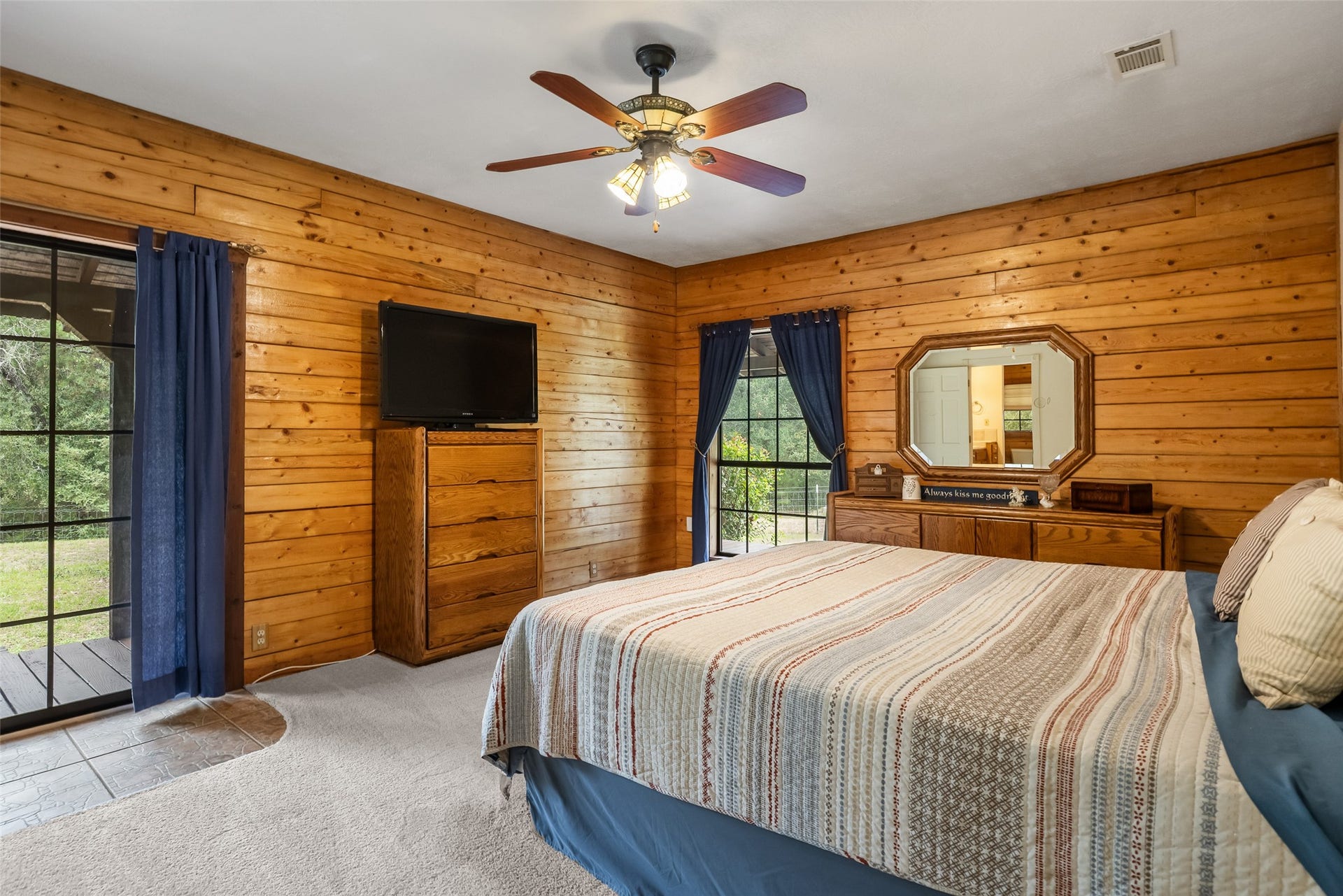 27874 Riley Road, Waller, TX 77484 The spacious first floor primary suite offers a comfortable retreat with warm wood walls, soft carpet, a cooling ceiling fan and large windows overlooking the peaceful countryside.
