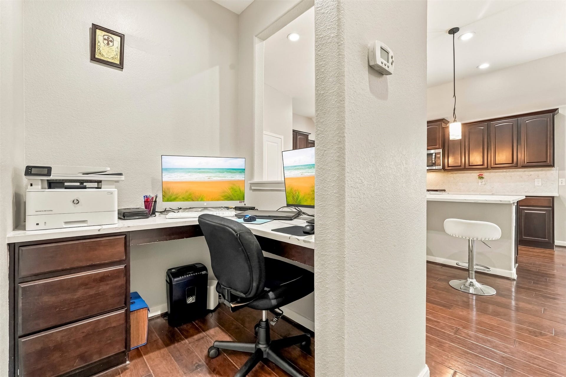 4557 Argonne Woods Drive, Porter, TX 77365 Modern home office nook with built-in desk, ample storage, and dual monitors, seamlessly connected to a stylish kitchen with dark cabinetry and a breakfast bar.