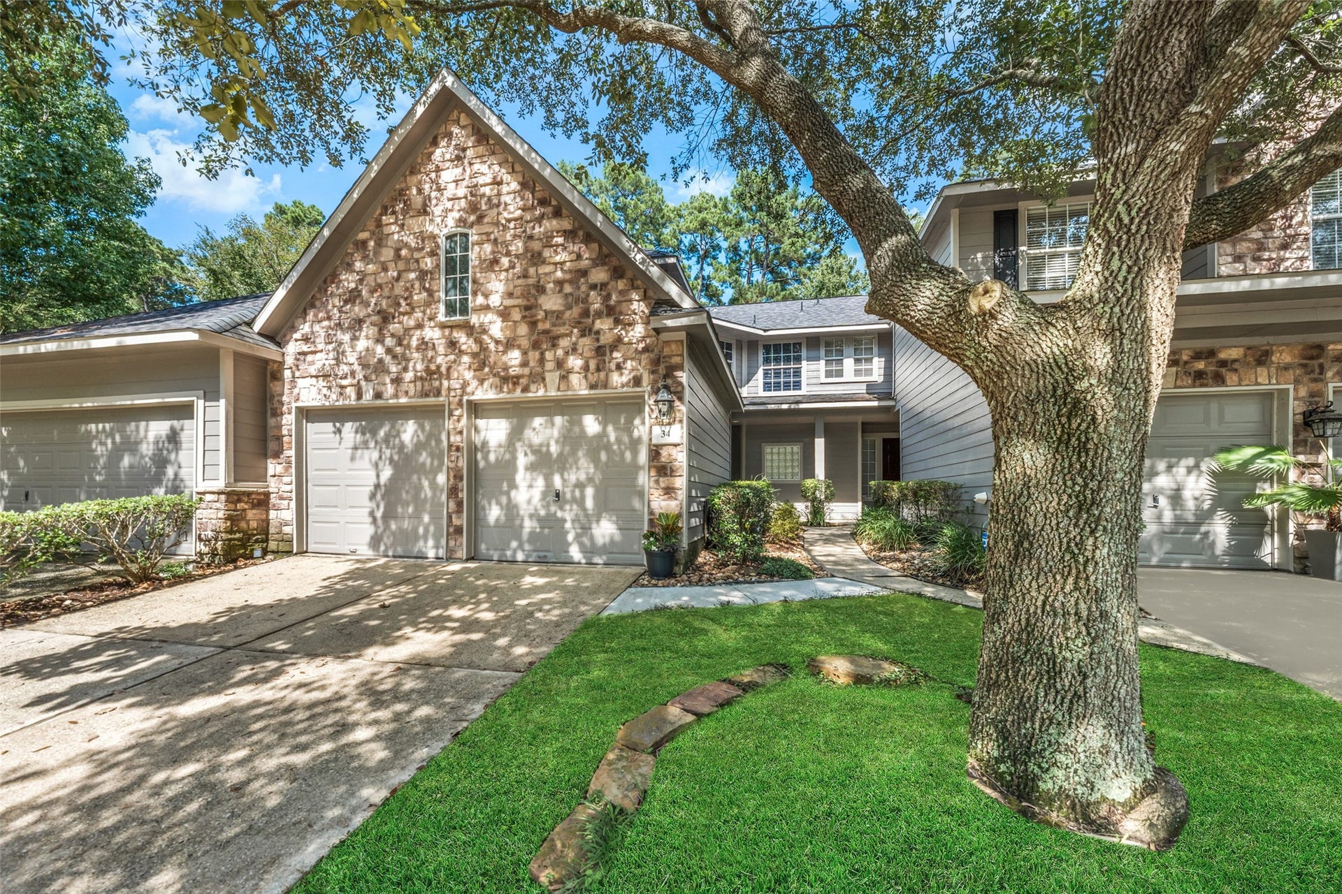 34 Alderon Woods Place, The Woodlands, TX 77382 Charming townhouse featuring a stone facade and dual garage, nestled under mature trees with a well-maintained lawn.