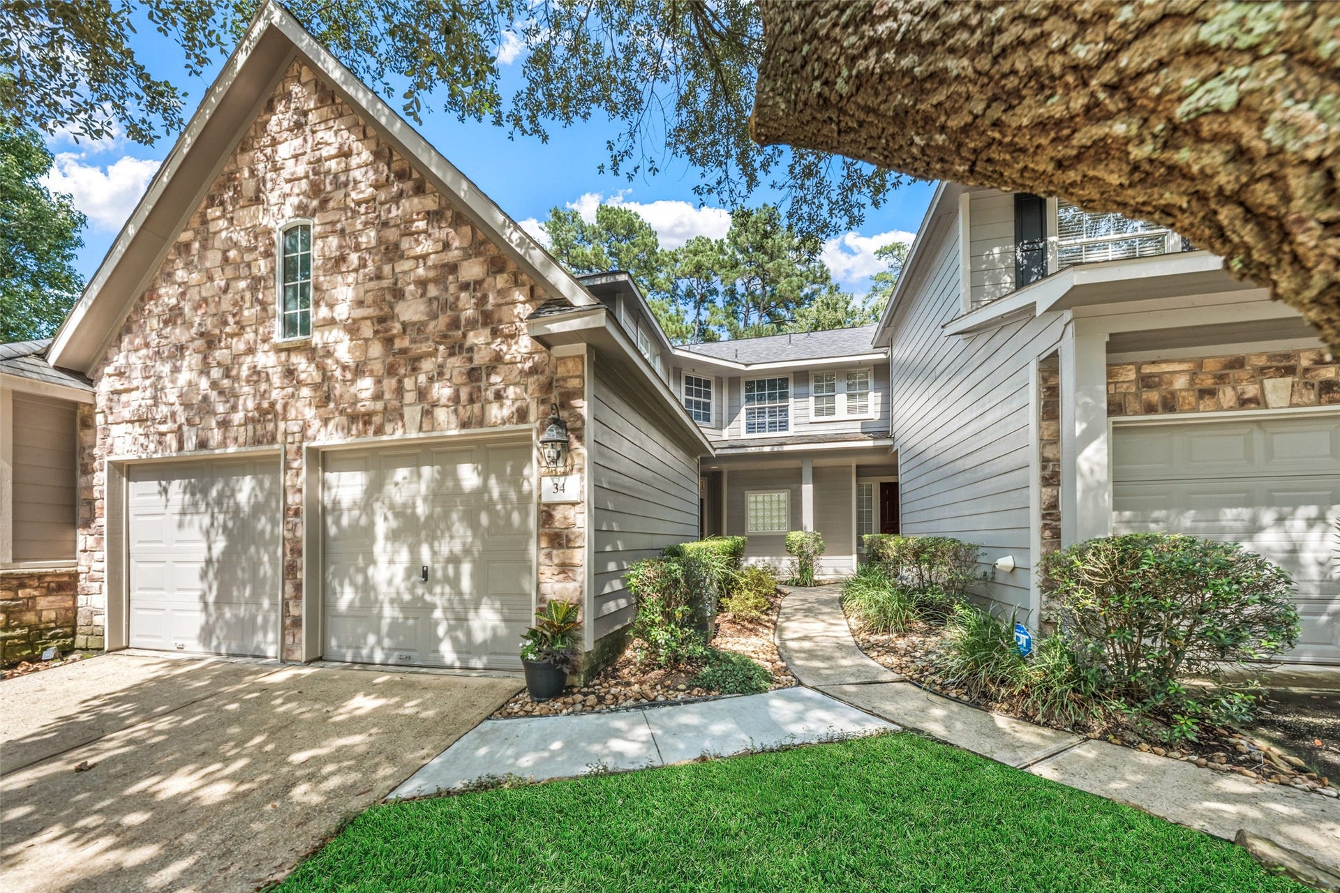 34 Alderon Woods Place, The Woodlands, TX 77382 Charming two-story home with a stone faade and two-car garage, nestled under mature trees, features a welcoming pathway leading to the front entrance.