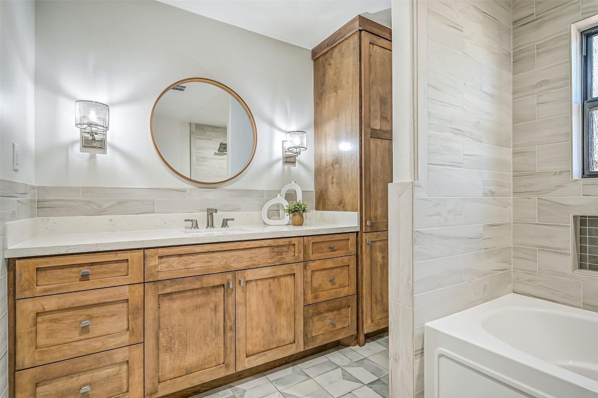 921 Falling Leaf Drive, Friendswood, TX 77546 The stylish guest bath showcases a quartz vanity with abundant stained cabinetry, tall linen storage, designer geometric tile flooring, contemporary sconces & a round wood-framed mirror, continuing the warm contemporary finishes found throughout the home.