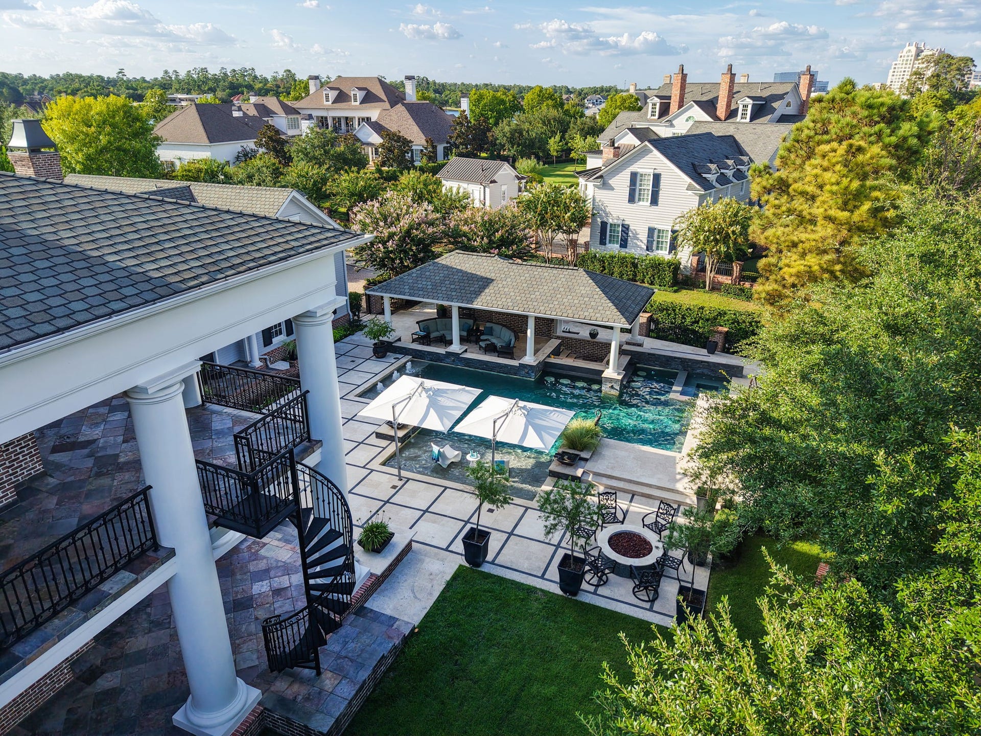 2 Georgian Row, The Woodlands, TX 77380 A breathtaking aerial perspective captures the full scale of this remarkable outdoor retreat. The resort-style pool serves as the centerpiece, surrounded by expansive entertaining spaces, a covered pavilion, manicured grounds, and the homes impressive columned veranda with a sweeping spiral staircase connecting the upper and lower outdoor spaces.