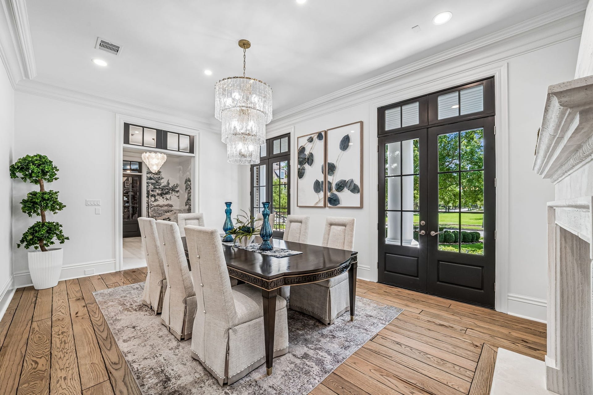 2 Georgian Row, The Woodlands, TX 77380 Elegant formal dining room featuring a statement crystal chandelier, fireplace, rich hardwood flooring, and French doors opening to the veranda and landscaped grounds.