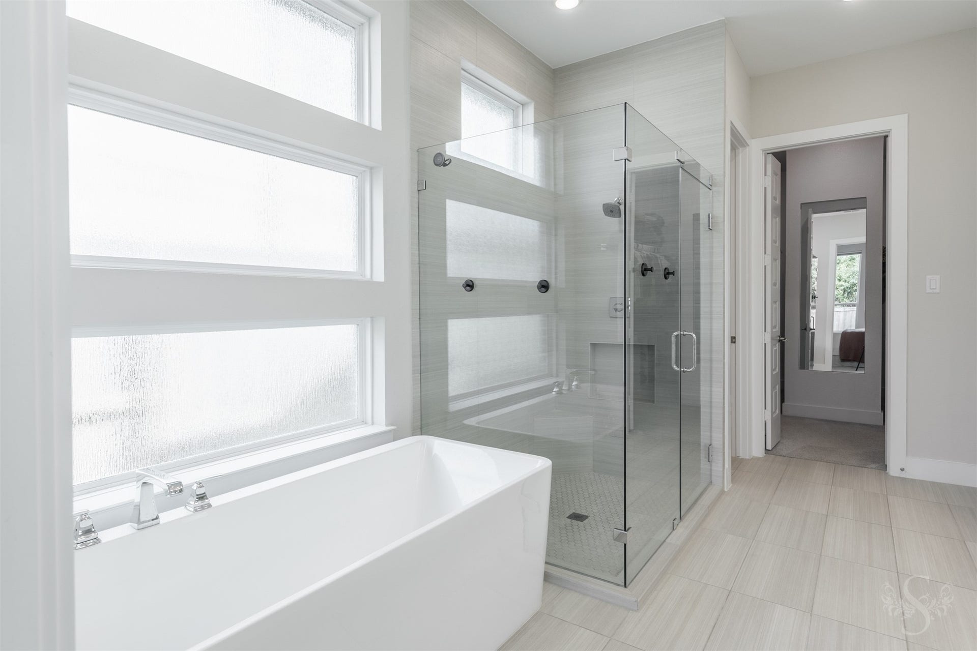 Freestanding soaking tub rests beneath a striking arrangement of frosted windows for privacy and natural light. An oversized walk-in shower with floor-to-ceiling tile and frameless glass creates a stunning companion feature.