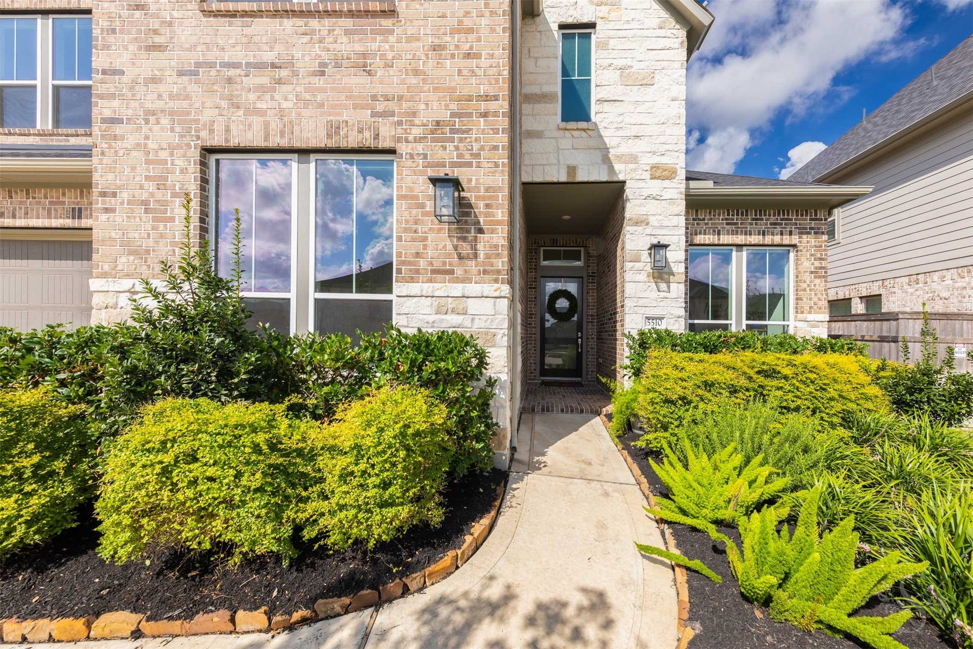 Lush, manicured landscaping and handsome brick-and-stone architecture frame the covered entrance. Contemporary exterior lighting and a modern glass-paneled door offer a sophisticated welcome.