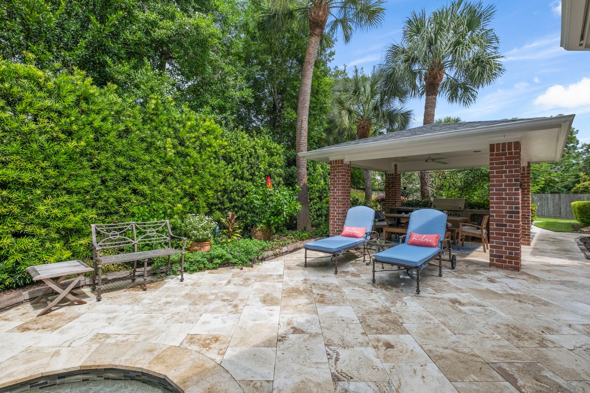 7 Silverloch Court, Sugar Land, TX 77479 A beautifully landscaped backyard features a spacious patio with elegant travertine flooring, space for a cozy seating area, and a charming brick pavilion perfect for outdoor dining. Lush greenery and palm trees provide a serene and private oasis.