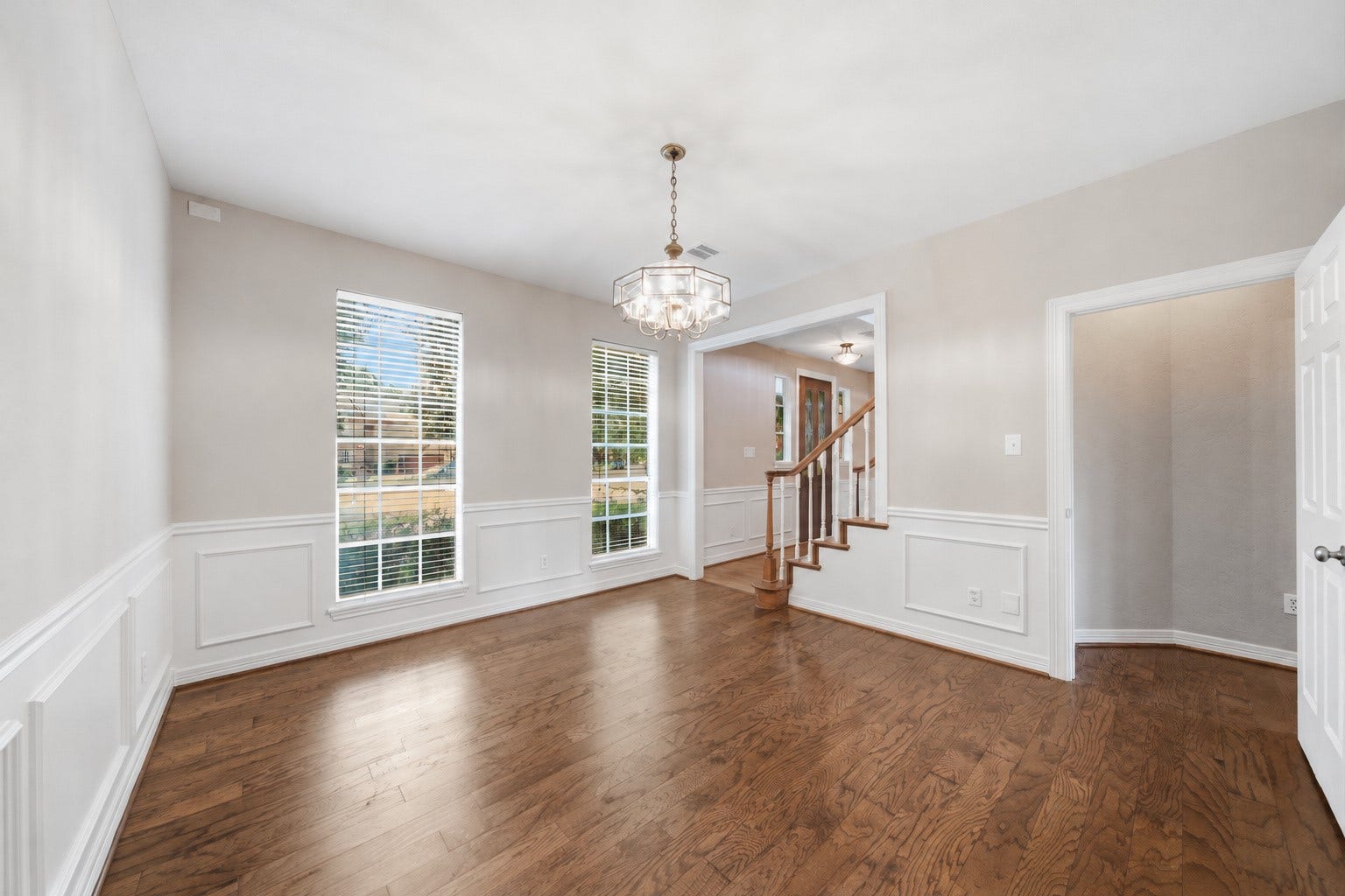 Elegant flex area for dining, office or lounge with rich hardwood floors, decorative wainscoting, and fresh paint leading to a staircase with wooden accents.