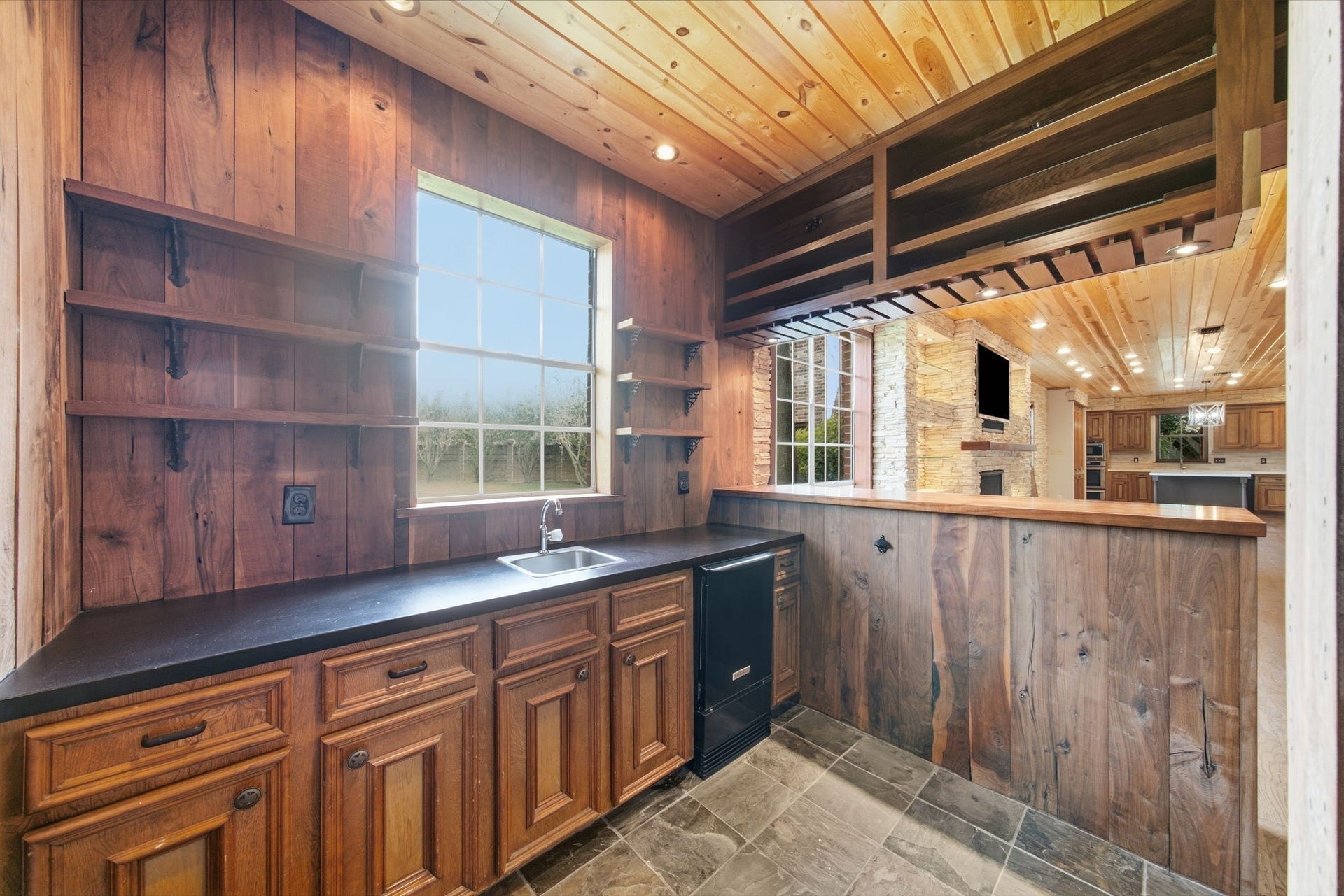 Rustic wet bar with rich wood paneling, sleek countertops, and ample shelving, featuring wine storage, sink, wine fridge and TV outlet. Seamlessly connects to a cozy living area with stone fireplace.
