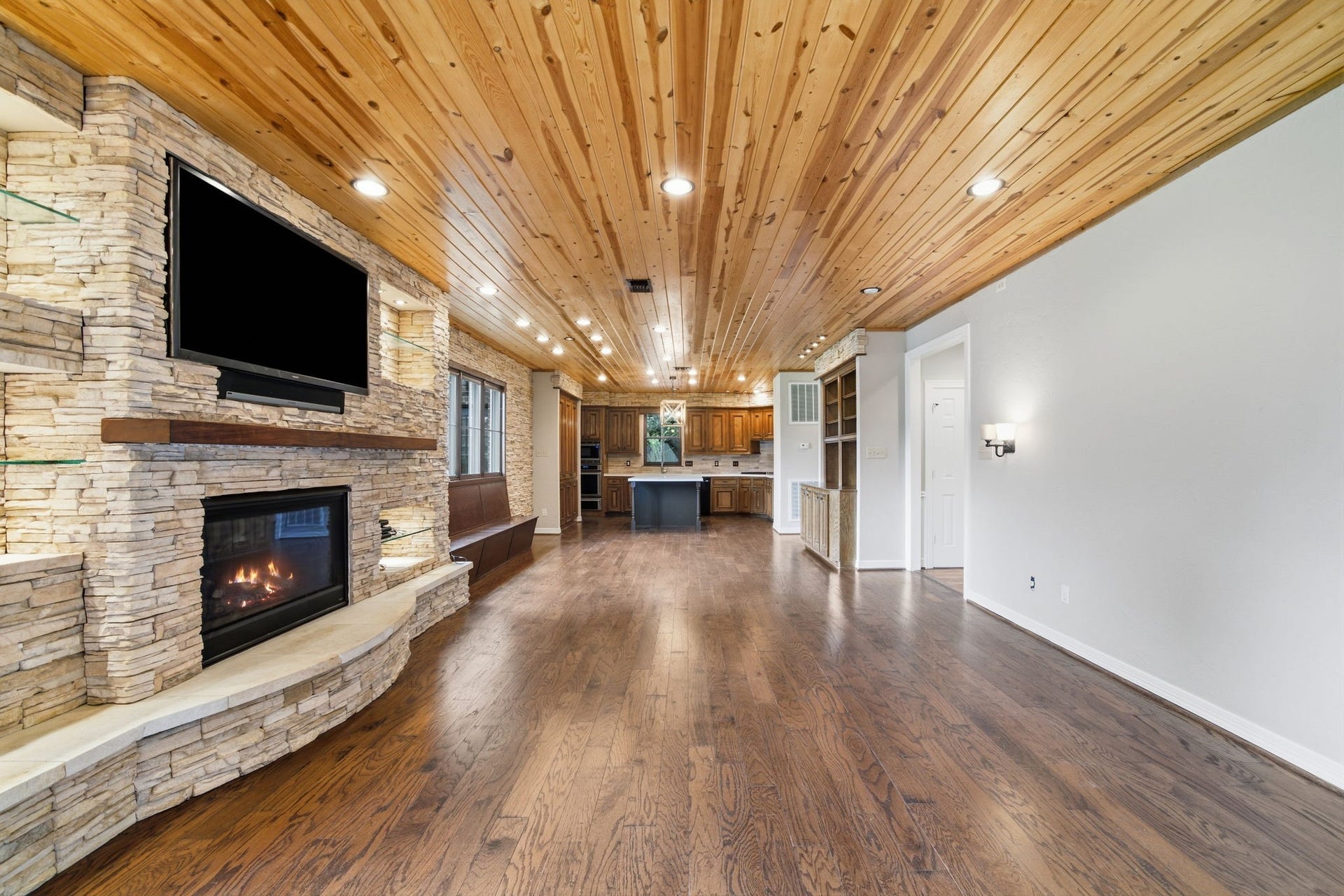 Stunning living area featuring a cozy stone fireplace with built-in TV, rich wooden floors, and a warm wood-paneled ceiling, seamlessly flowing into an inviting kitchen space.