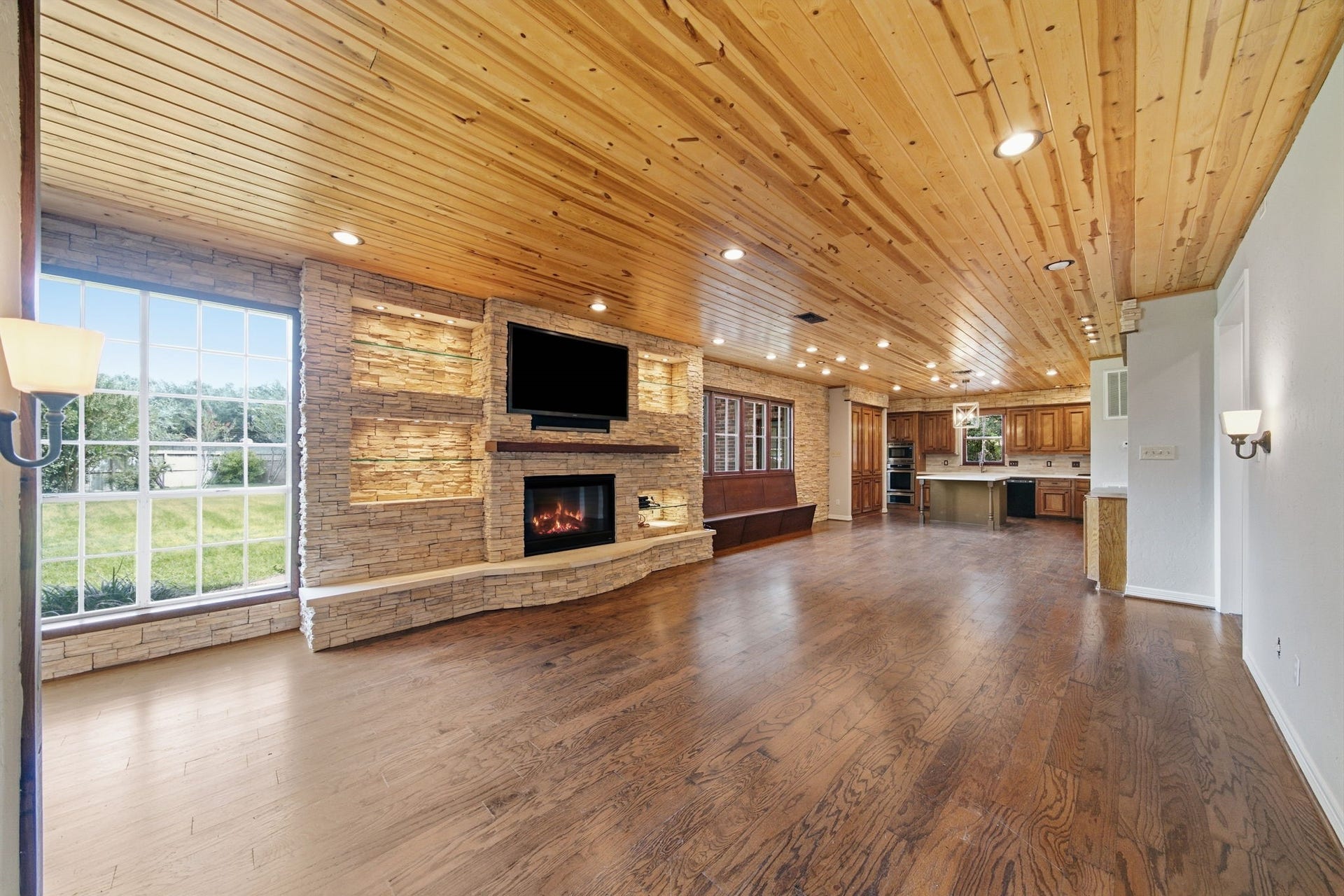 Spacious open-concept living area featuring a cozy fireplace with stone accents, rich wood flooring, and a warm wood ceiling, seamlessly flowing into the modern kitchen. Expansive windows provide abundant natural light and scenic views.