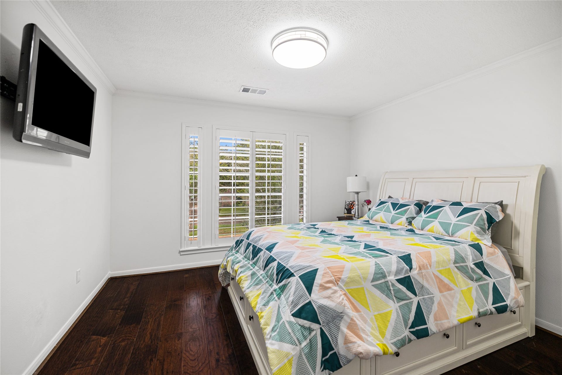 Spacious secondary bedroom featuring engineered wood flooring, crown molding, and plantation shutters overlooking the front yard.