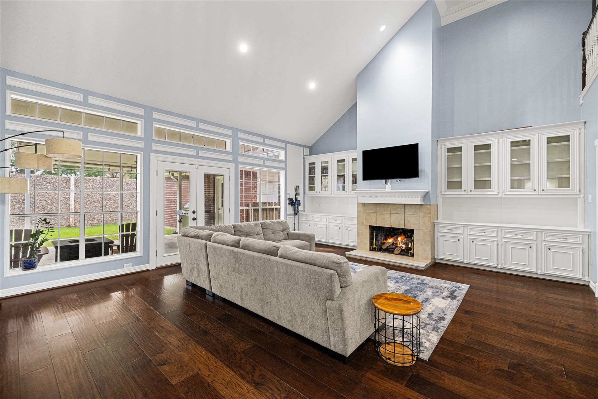 Spacious two-story family room featuring soaring ceilings, rich wood flooring, cozy fireplace, and a wall of windows overlooking the backyard.