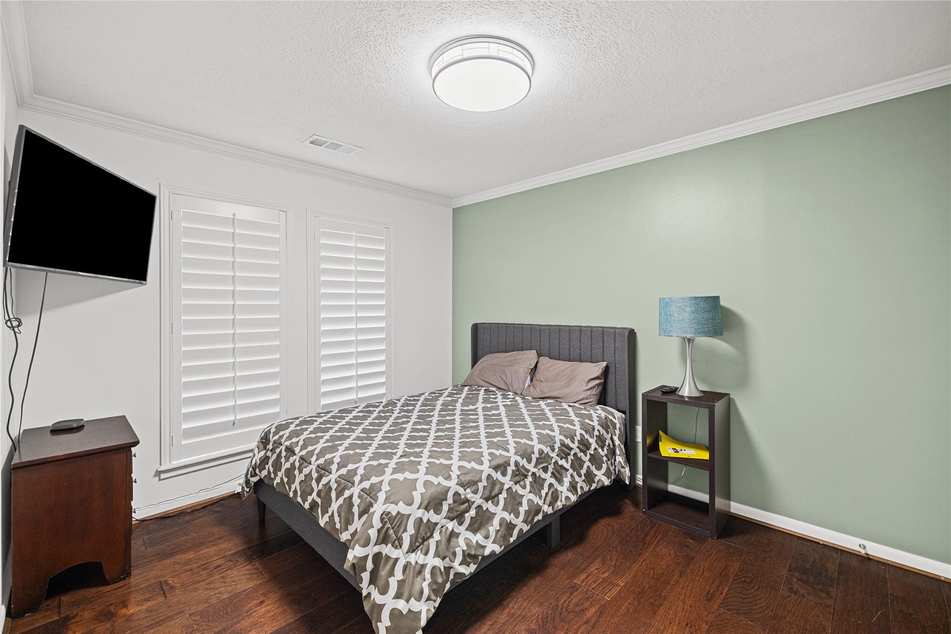 Bright secondary bedroom featuring engineered wood flooring, plantation shutters, and a neutral accent wall.