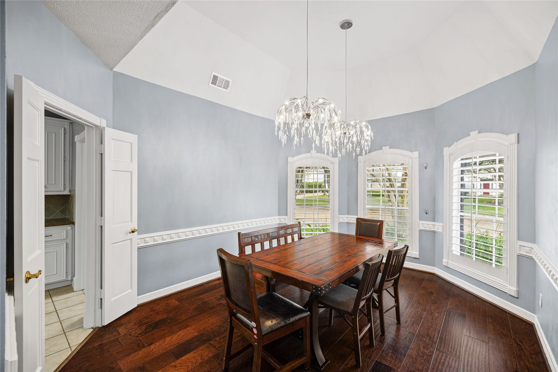 Elegant formal dining room with soaring ceiling, rich wood flooring, decorative chair rail molding, three arched windows with plantation shutters, and stunning chandeliers.