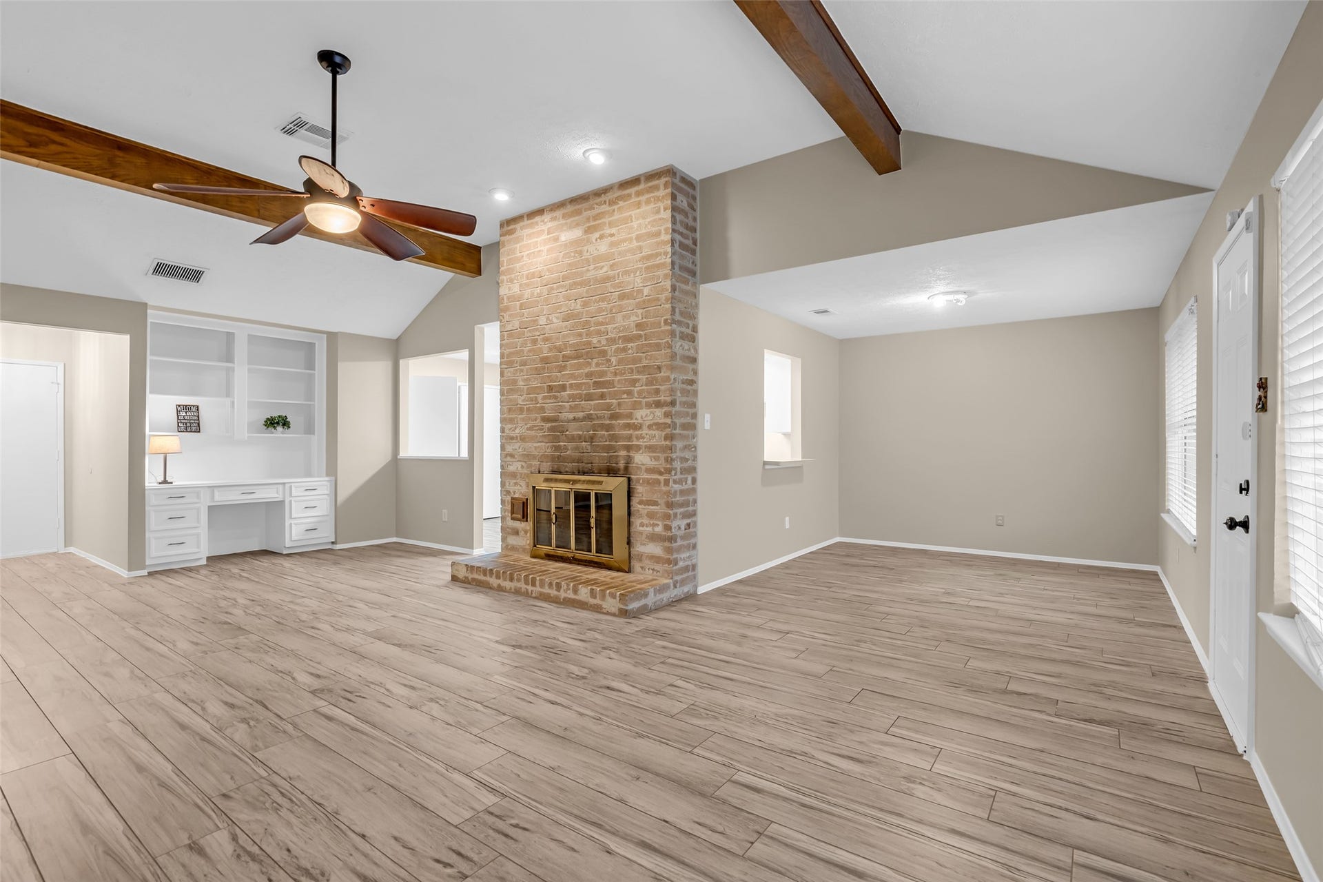 11827 Brook Meadows Lane, Meadows Place, TX 77477 This spacious living area features a vaulted ceiling with exposed wooden beams, a brick fireplace, and a ceiling fan. Built-in shelving with a desk for convenience and ample natural light from multiple windows.