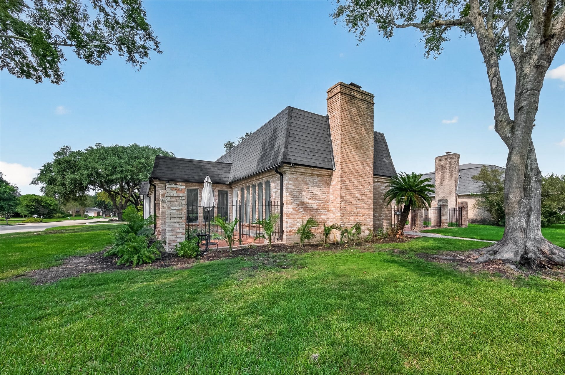 2230 S Fountain Valley Drive, Missouri City, TX 77459 Corner view of the property showing the brick chimney, sloped rooflines, enclosed patio area, palm landscaping, and mature trees along the front and side yard. The home sits on a green, tree-lined street with a broad lawn and established neighborhood