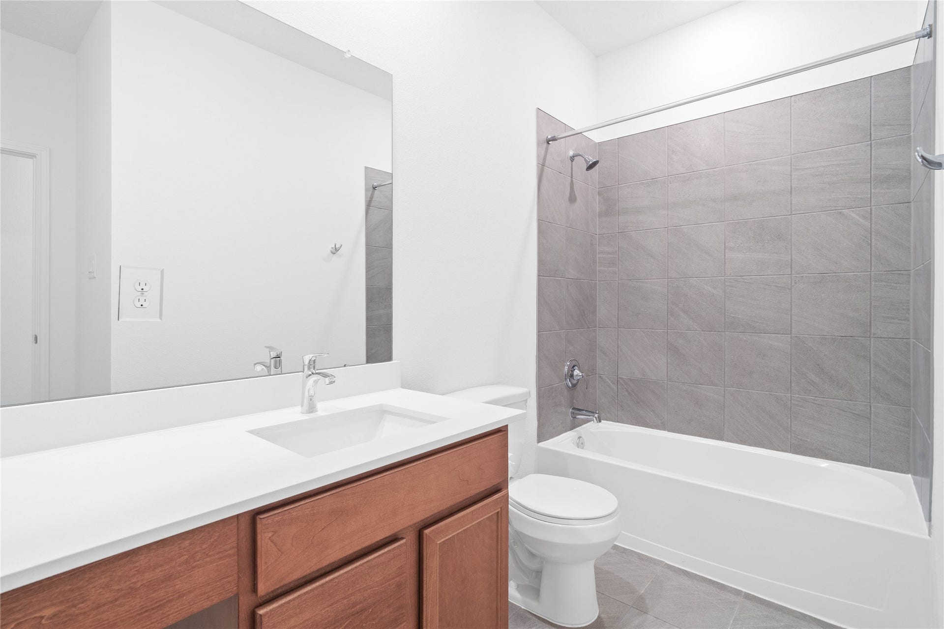 The secondary bathroom offers tile flooring, a bath/shower combination with a stylish tile surround, rich stained wood cabinetry with added knee space and elegant light countertops, a mirror, and sleek fixtures with modern finishes.