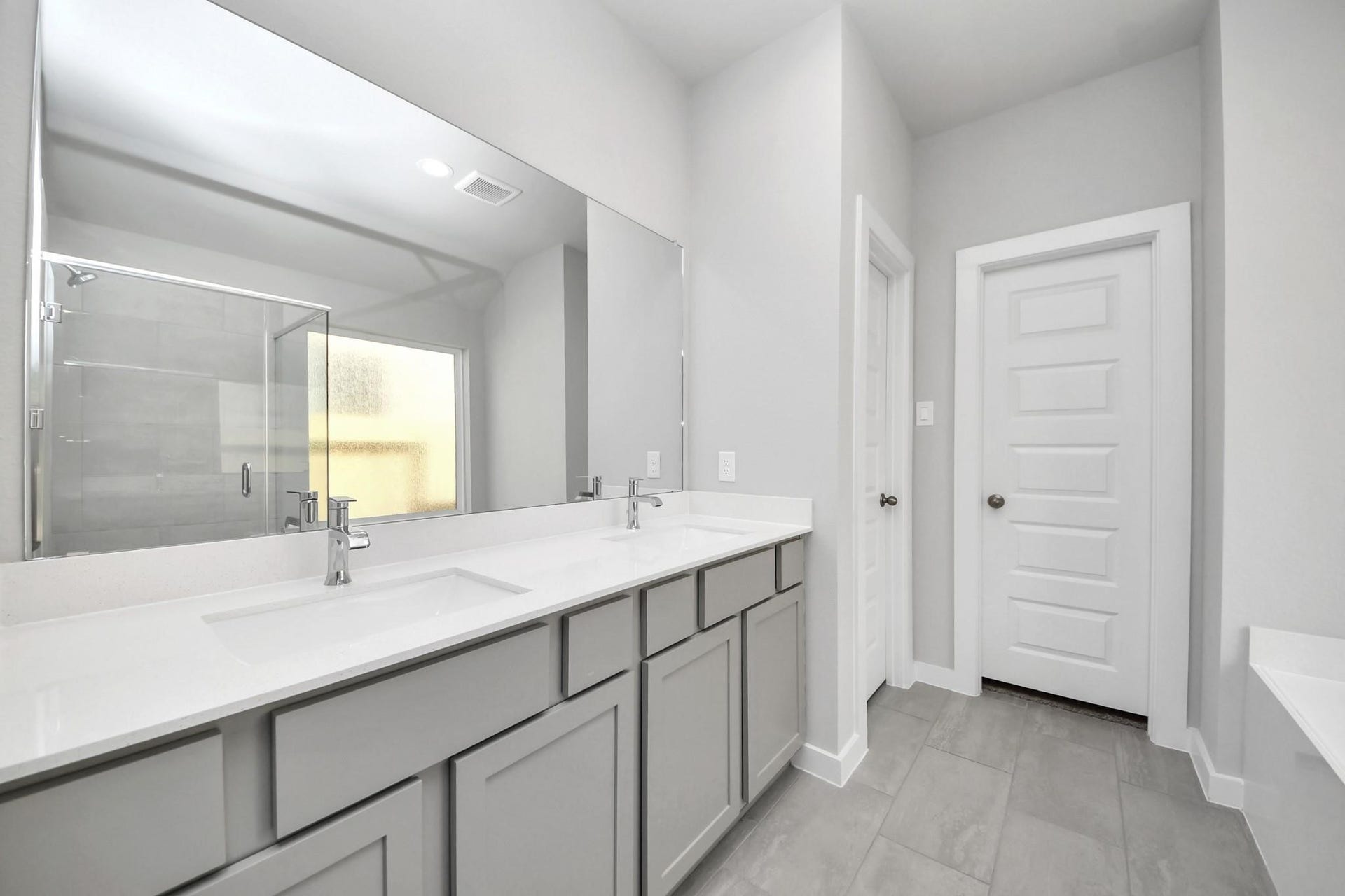 Spacious bathroom featuring a double vanity with sleek gray cabinetry, a large mirror, and modern fixtures, complemented by a glass-enclosed shower and elegant tile flooring.