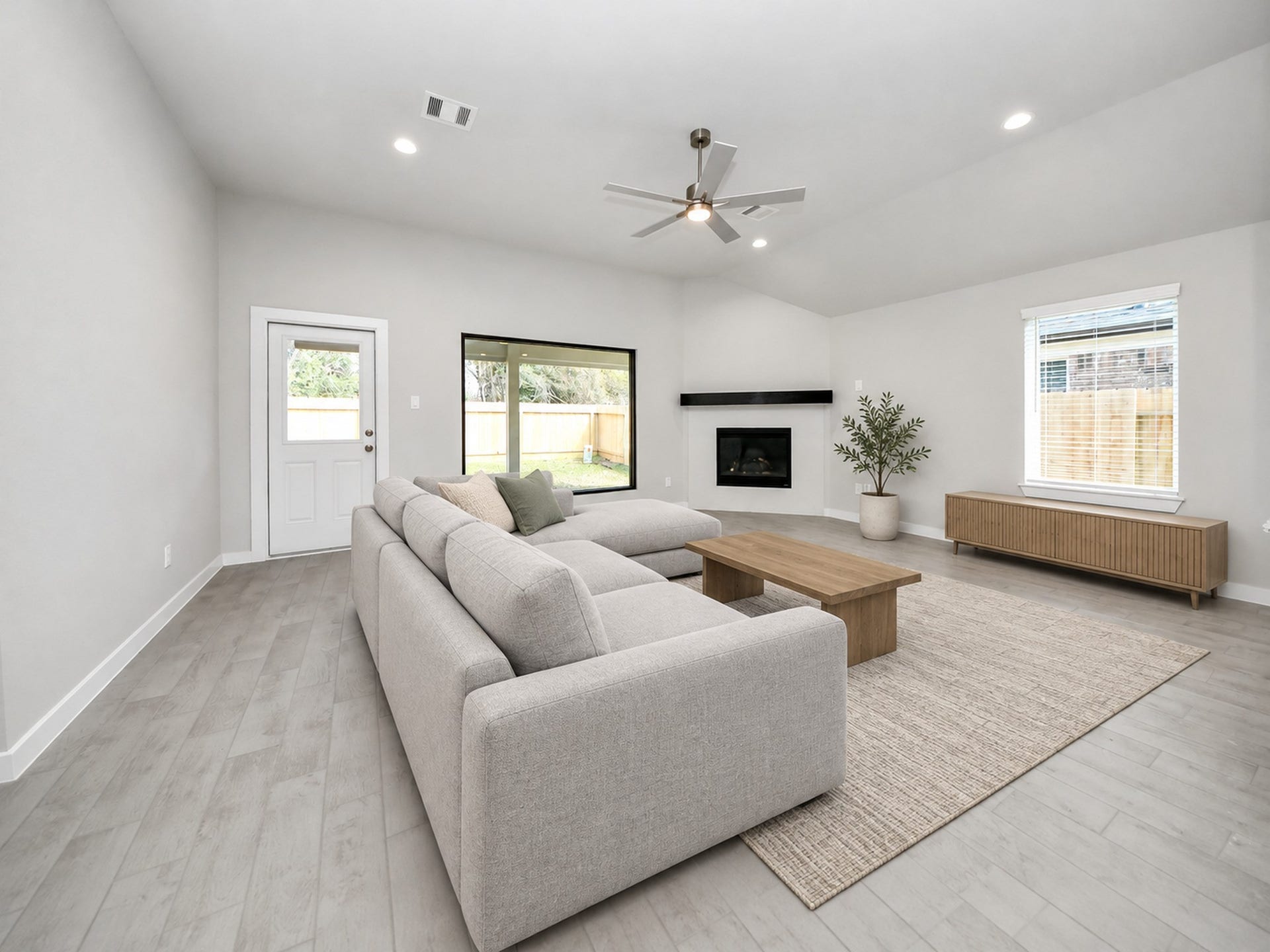 Spacious living room with modern gray flooring, large sectional sofa, and a sleek fireplace. Natural light pours in through a large window and glass door, enhancing the open and airy feel.