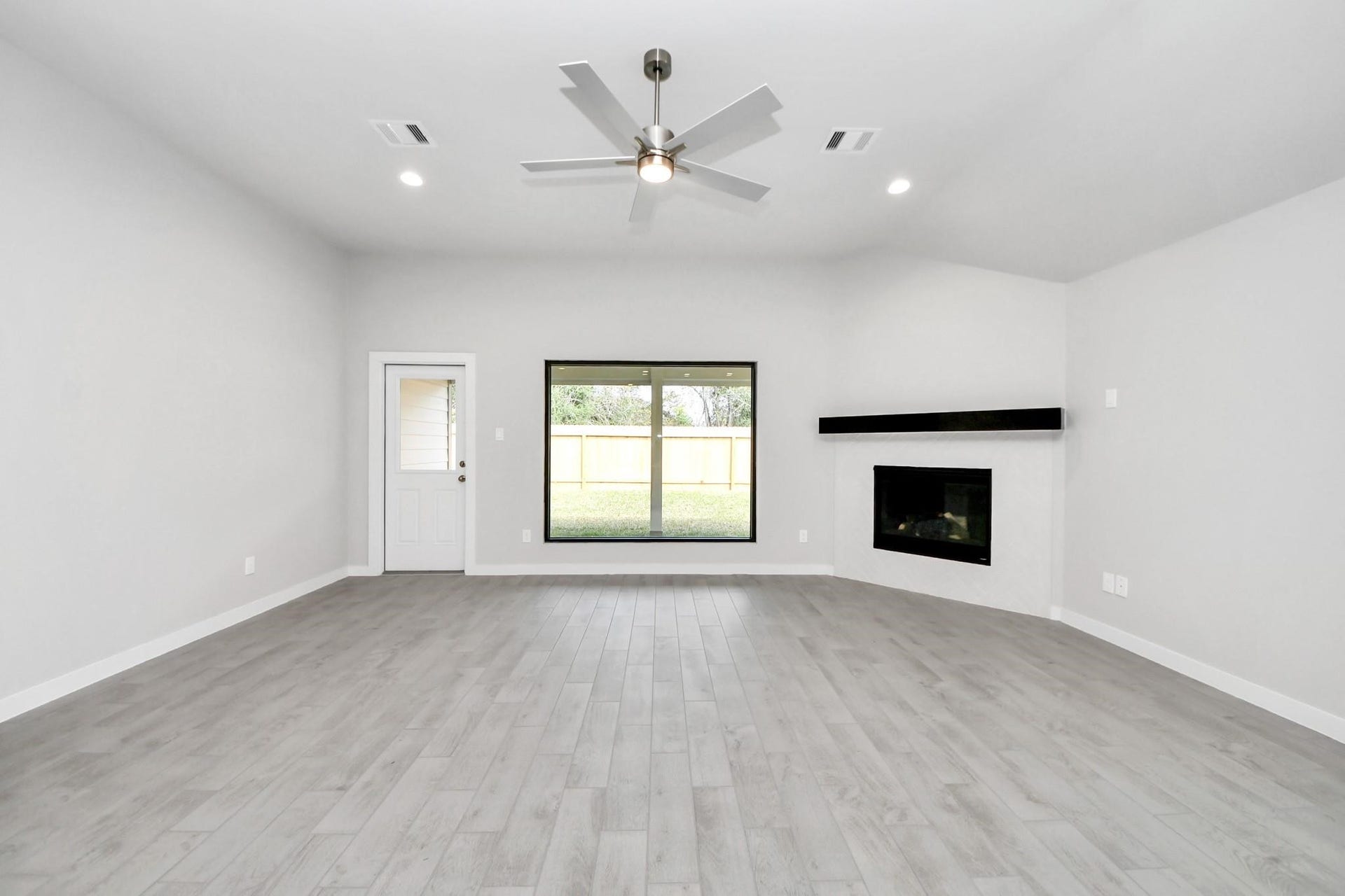 Spacious living room with sleek hardwood floors, a modern ceiling fan, and a cozy corner fireplace, all enhanced by ample natural light from a large window and convenient patio access.