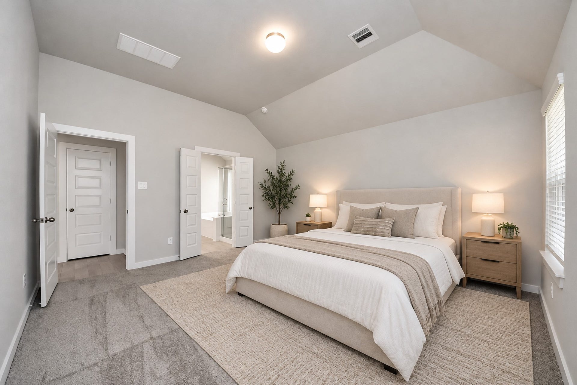 Spacious bedroom with vaulted ceiling, plush carpeting, and soft neutral tones. Features a cozy bed, stylish nightstands, and ample natural light. Double doors lead to an en-suite bathroom for added convenience.