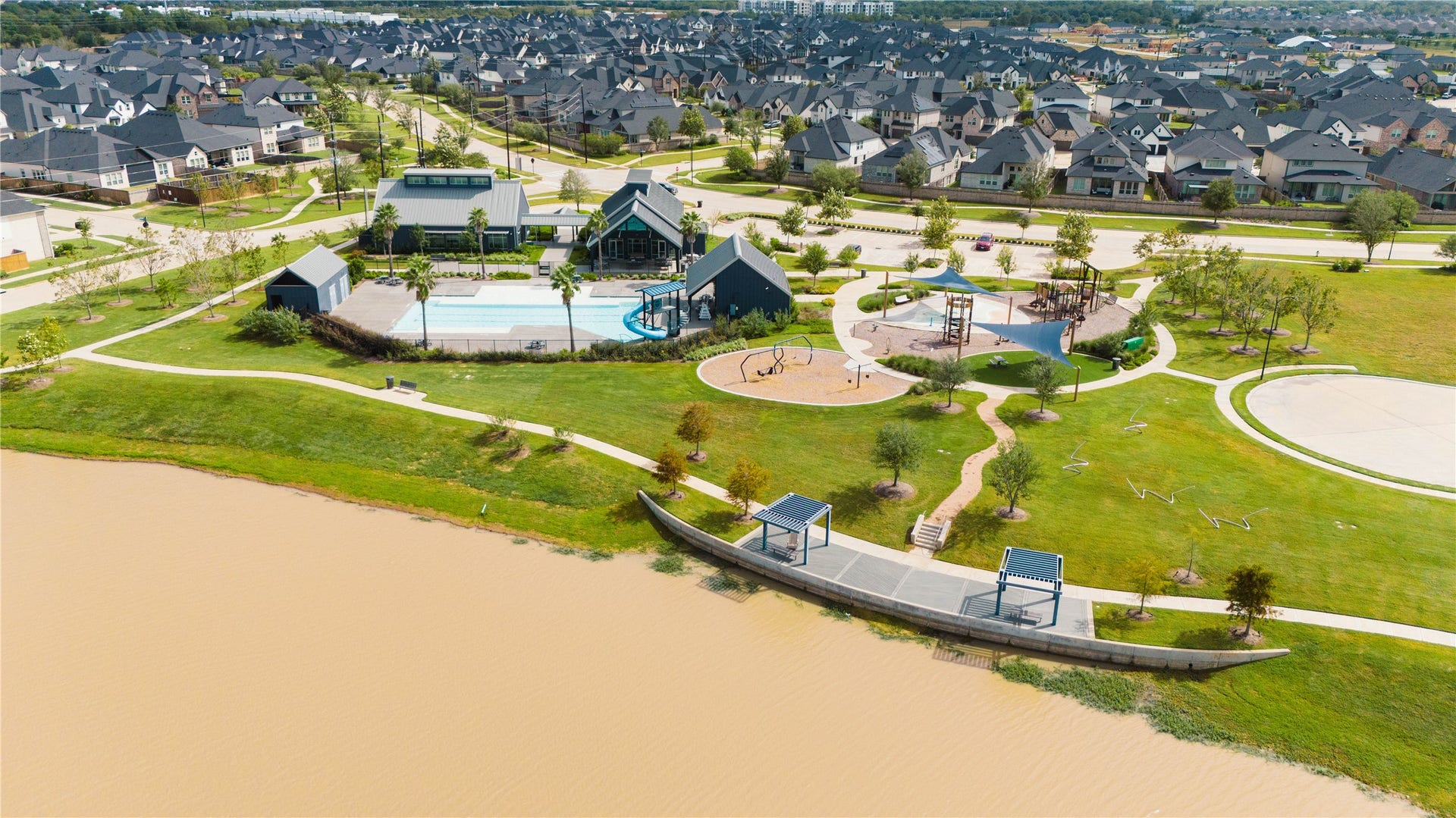 A breathtaking panoramic view of the Pecan Ridge amenity hub, highlighting the lakeside location with scenic shaded pavilions, winding walking paths, the community clubhouse, and a resort-style pool complex overlooking the water.