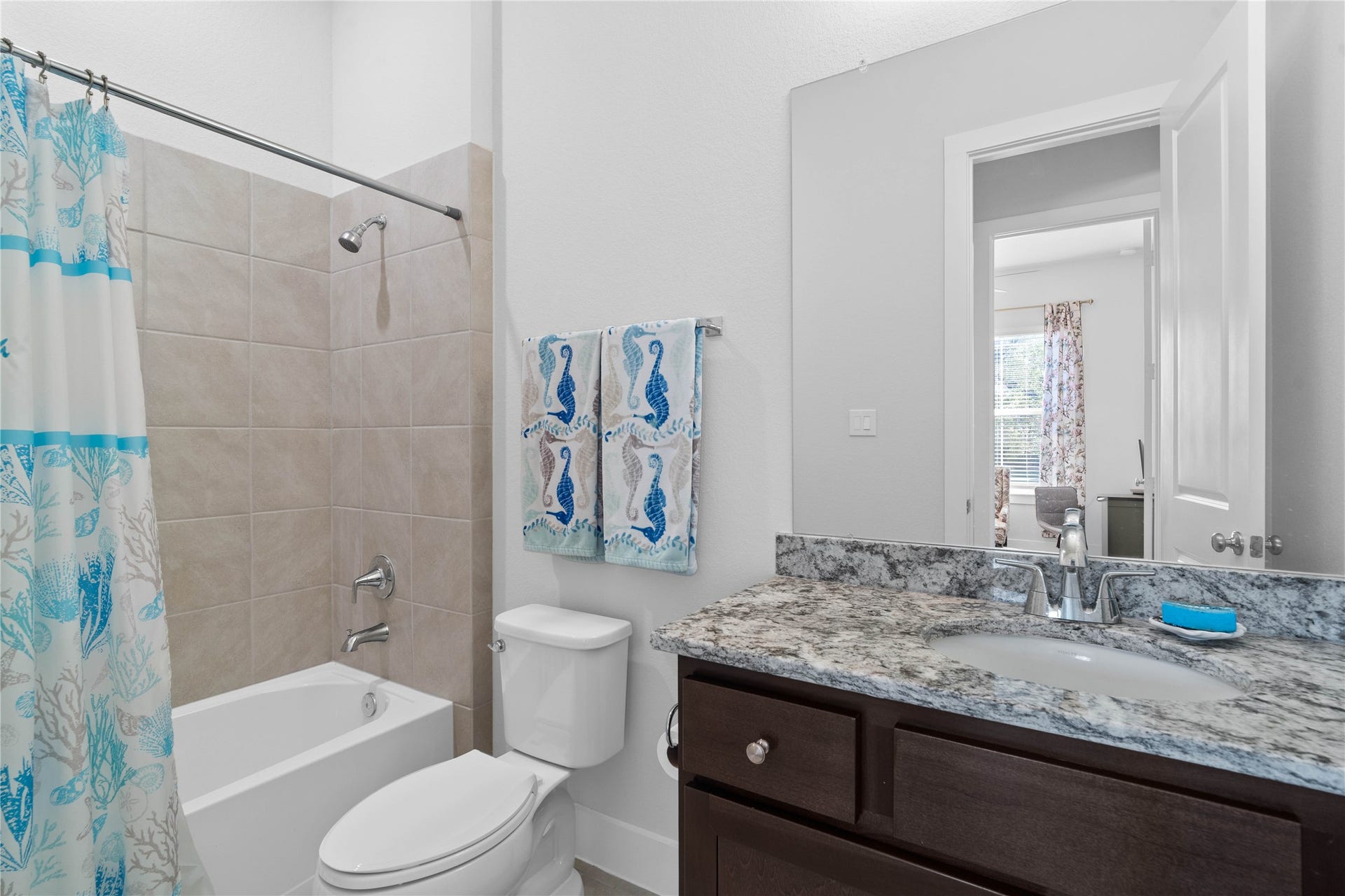 The secondary bathroom features a stunning granite vanity top, dark wood cabinetry, a large vanity mirror, and a  tub/shower combo accented by neutral wall tiling.