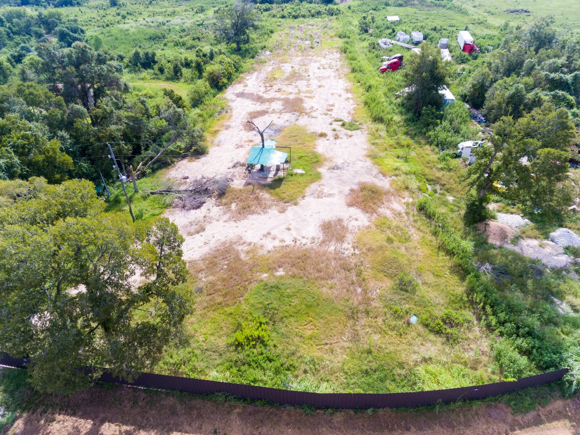 This aerial photo showcases a spacious, partially cleared lot surrounded by greenery, with a few scattered trees and some vehicles and structures in the distance. Ideal for development or personal projects, it offers a serene, natural setting with ample space.