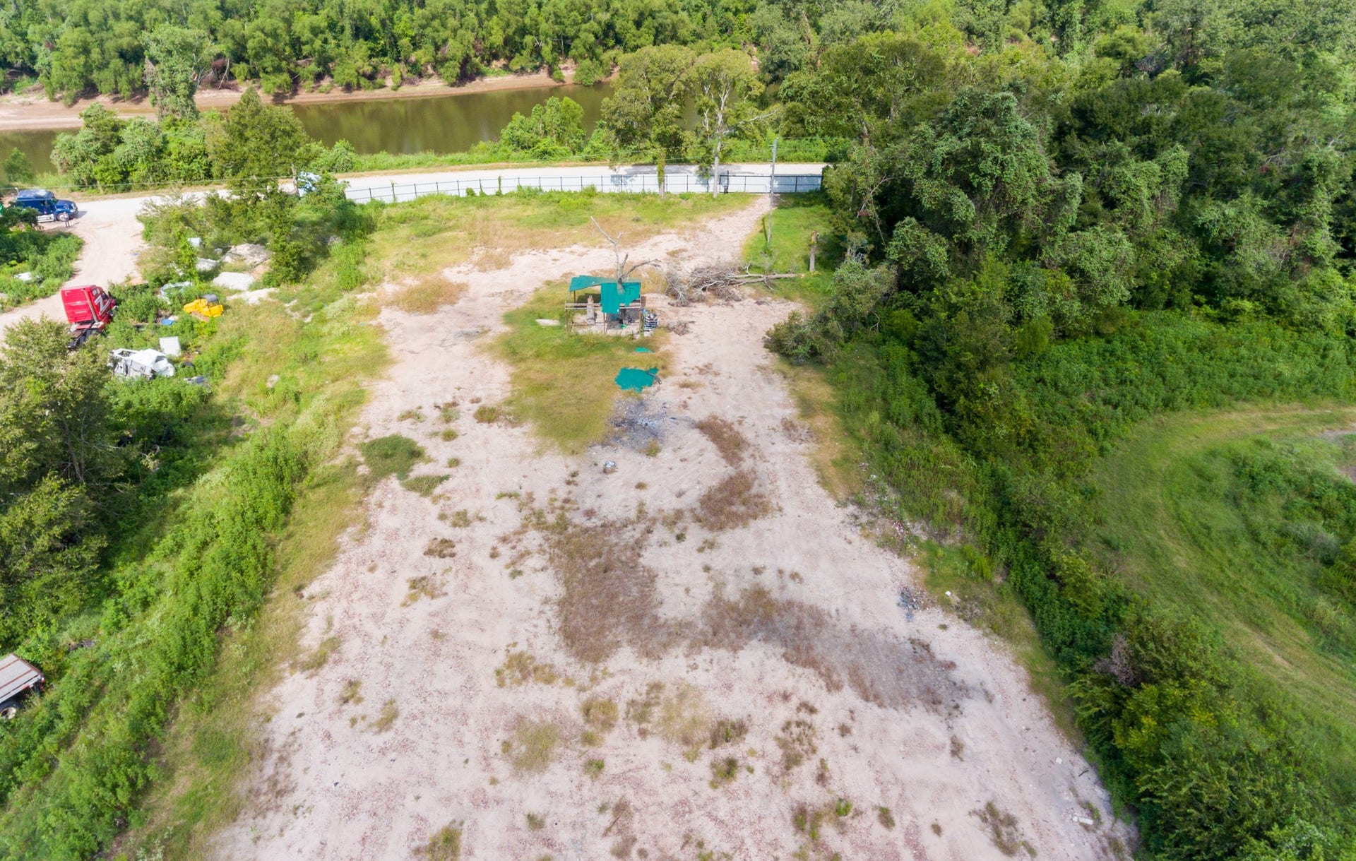 This photo shows a spacious, partially cleared lot with a mix of grassy and sandy areas, surrounded by trees. A road runs along one edge, and there's a view of a river in the distance, providing a natural and serene setting.
