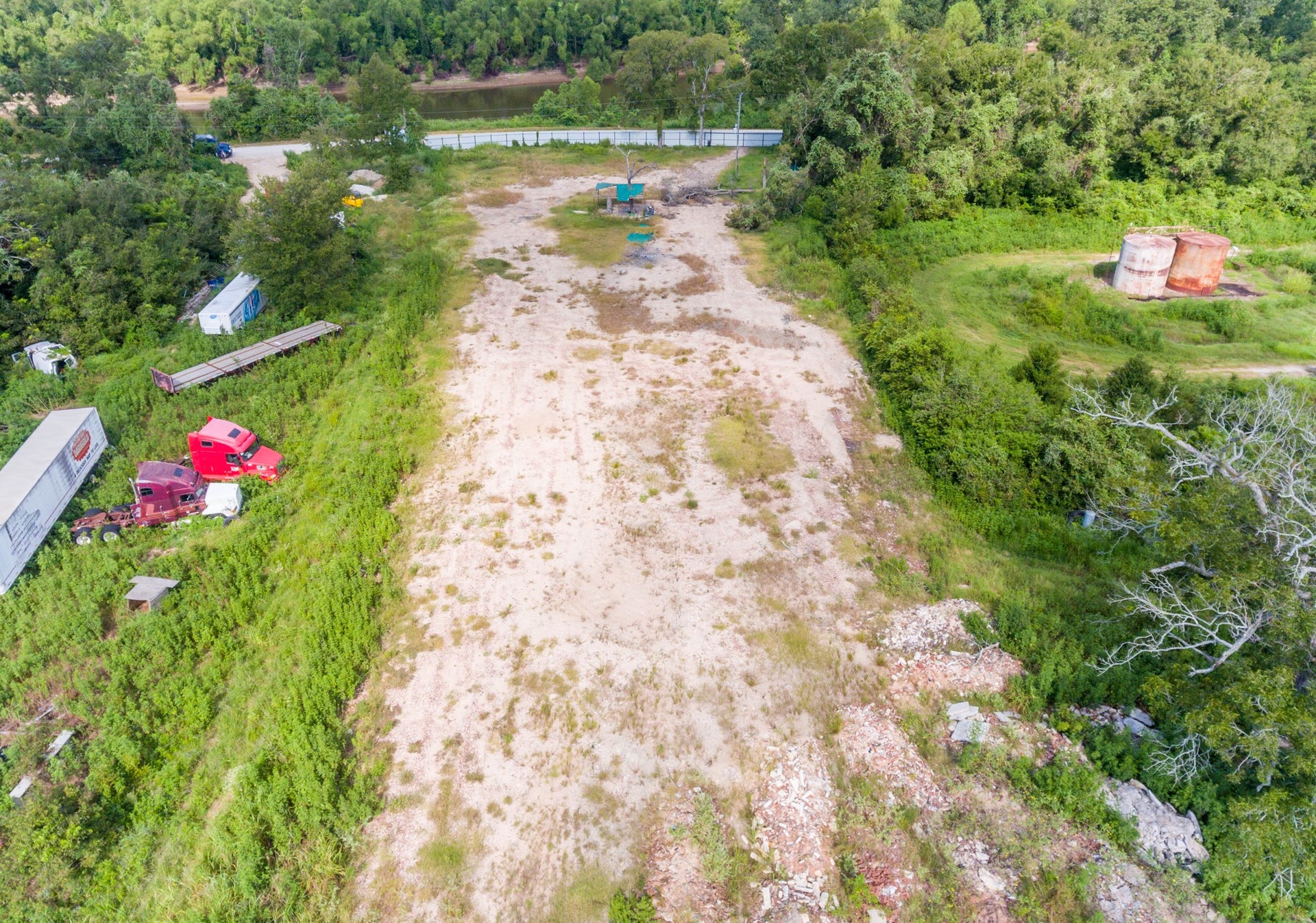 This photo shows a large, open lot with a mix of grassy and bare areas, bordered by trees. There are a few trucks and trailers parked on the left side, and an industrial tank is visible on the right. The lot appears to be near a road and surrounded by greenery.