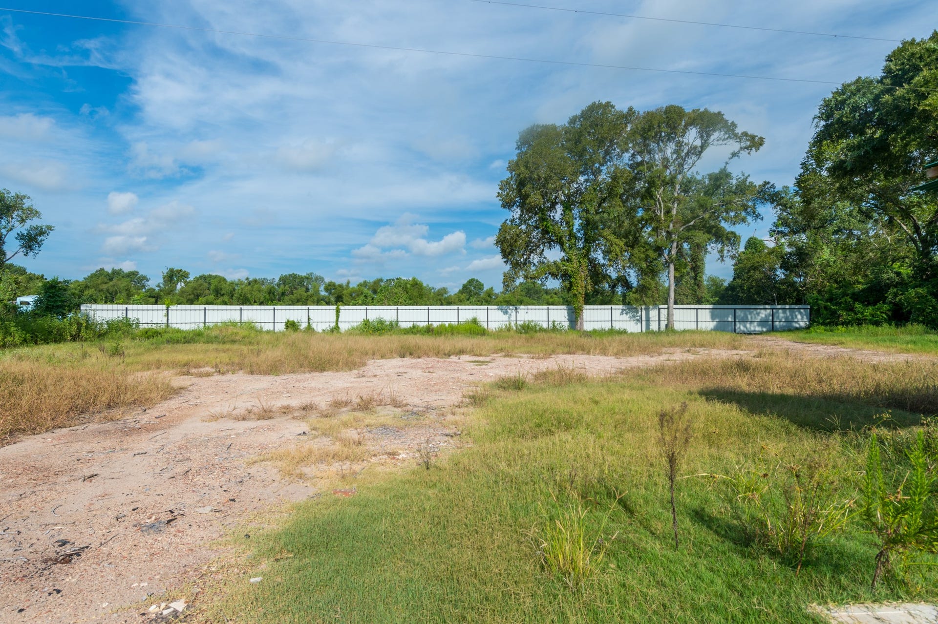 This photo shows a vacant plot of land with a mix of grass and dirt, bordered by a white fence and surrounded by trees, offering a natural setting with clear skies.
