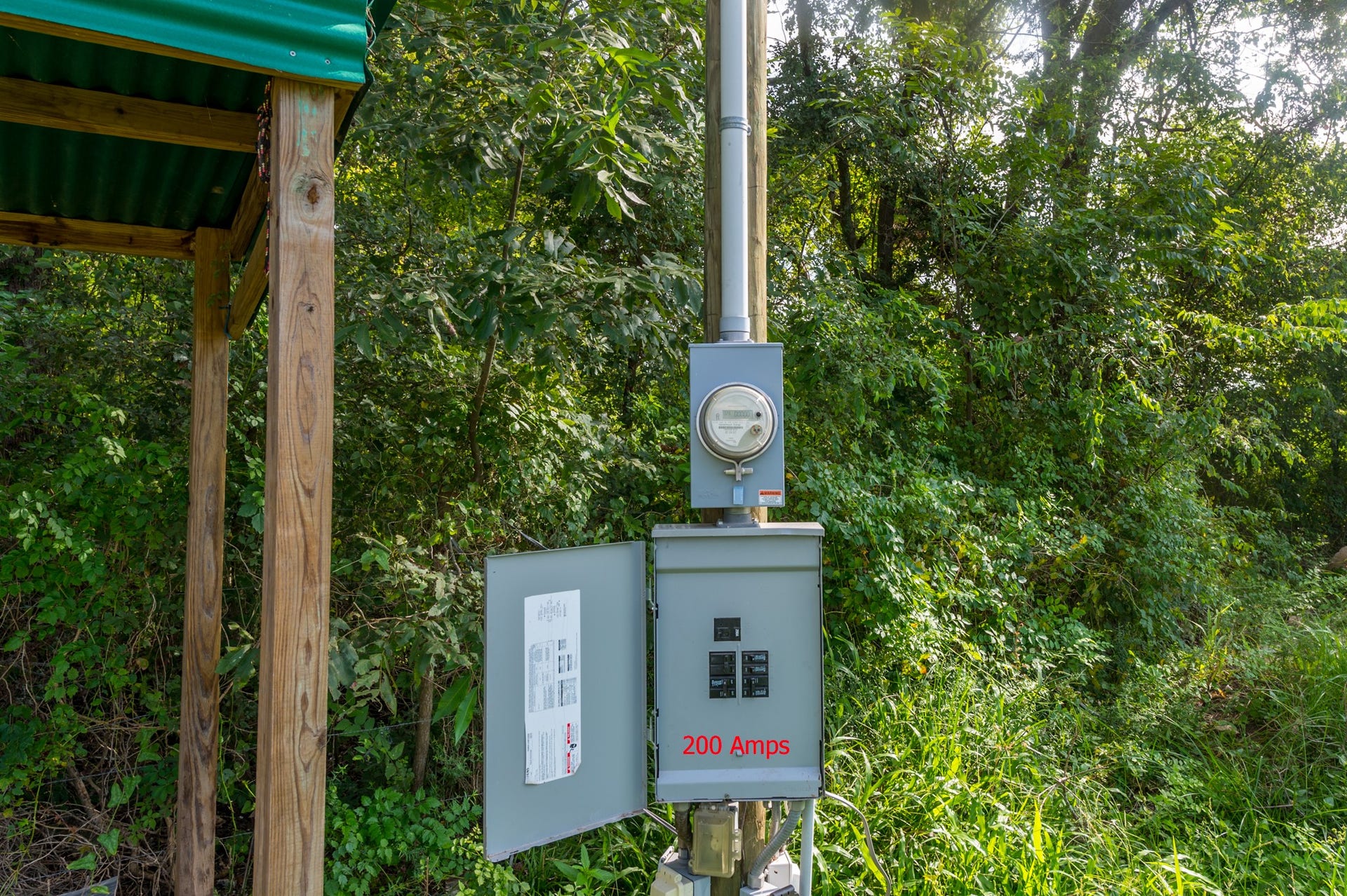 This photo shows an outdoor electrical meter and panel with 200 amps capacity, set in a lush, wooded area. The panel is mounted on a structure with a green roof for protection.