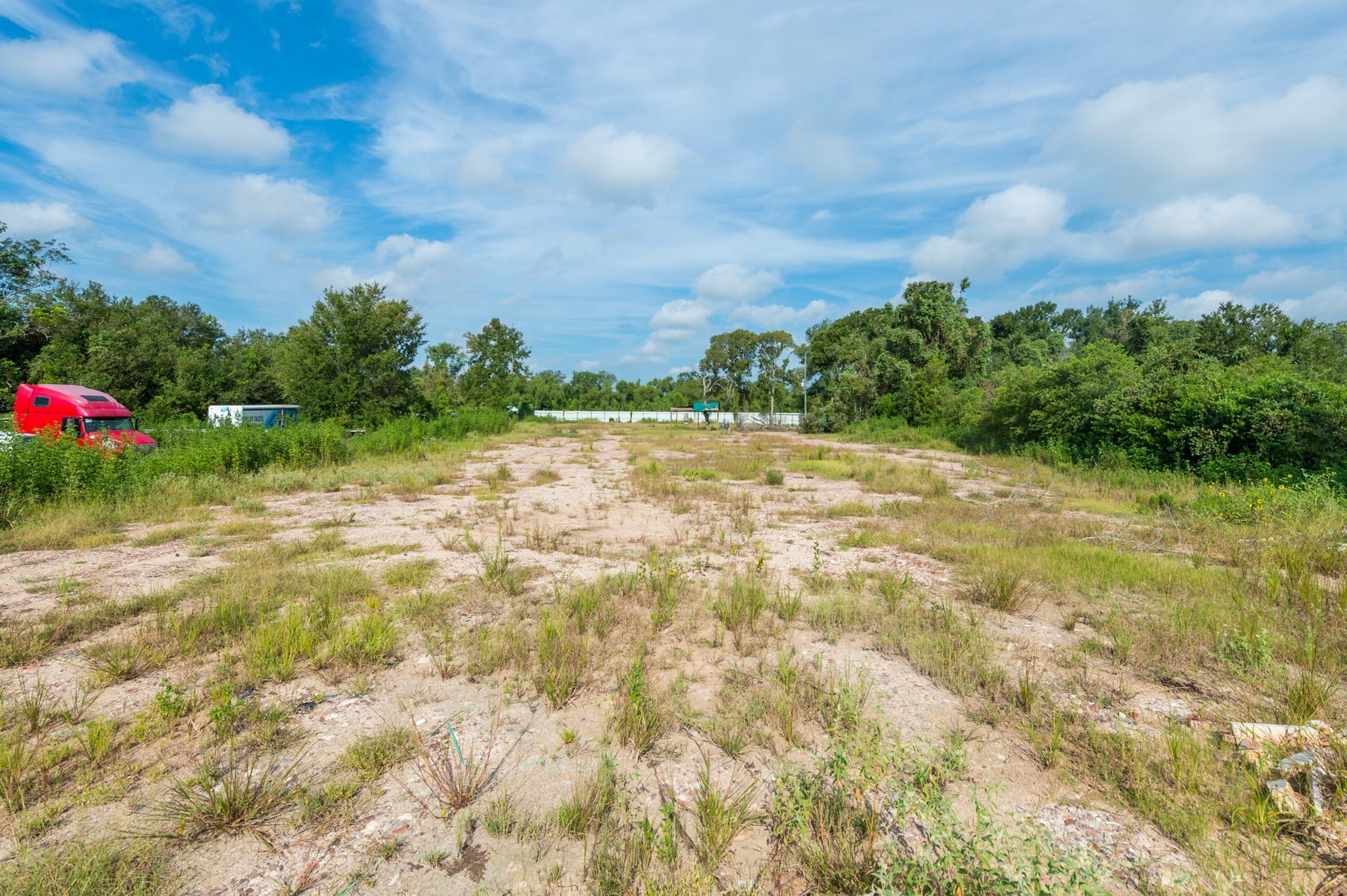 This photo shows a large, open plot of land with sparse vegetation and surrounding trees under a blue sky. Ideal for development or agriculture, it offers a spacious and natural setting.