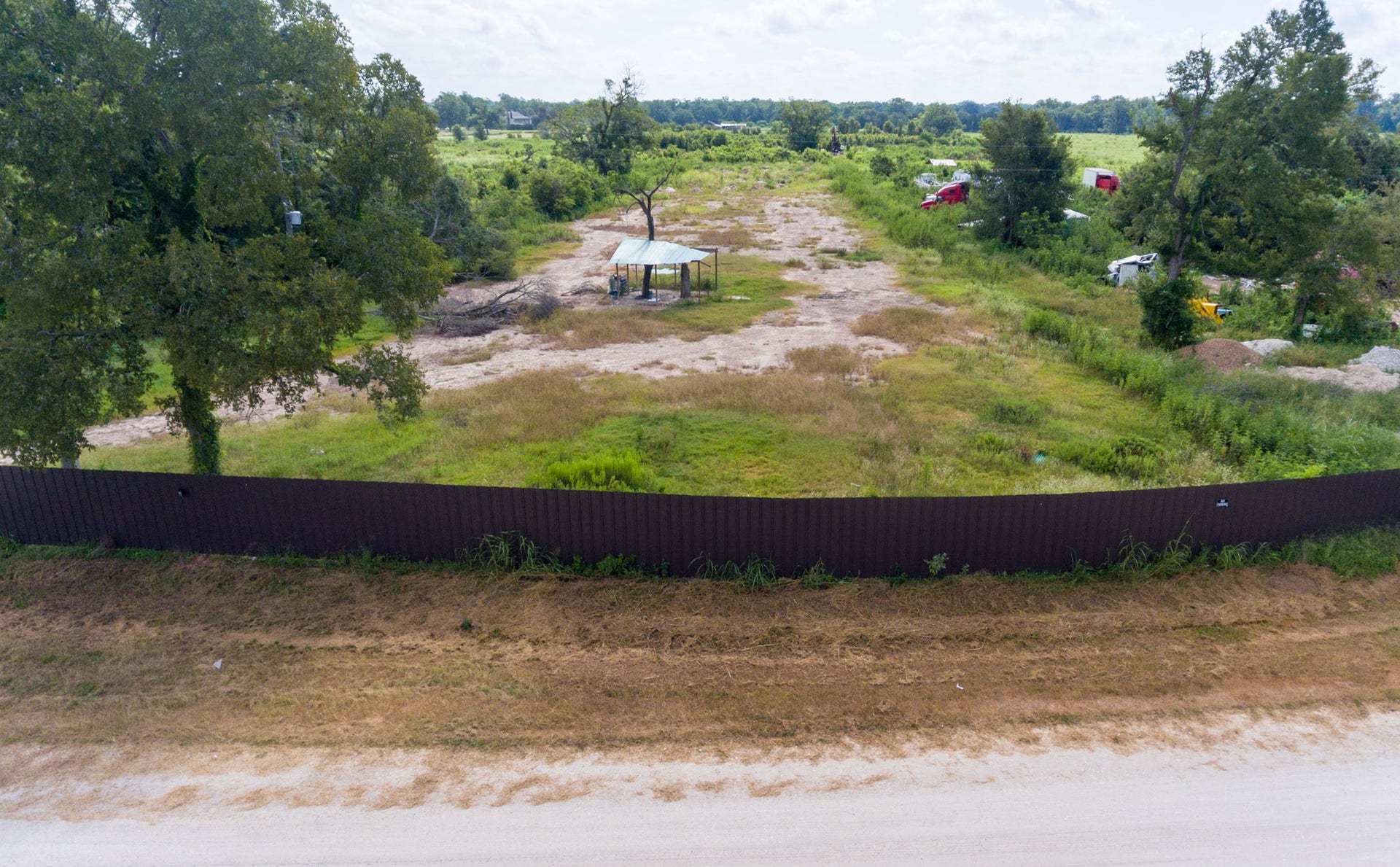 This photo shows a spacious, partially cleared lot with some trees and a small structure, surrounded by a fence. Ideal for development, it offers open land with a rural feel, and is accessible from a dirt road.