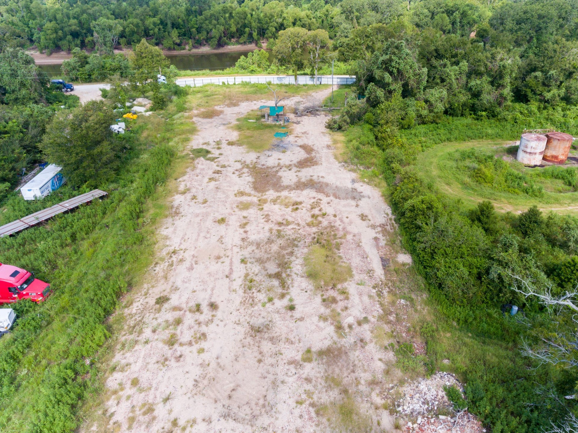 This photo shows a large, open plot of land surrounded by greenery. It features some sparse vegetation and an unpaved area, with a few vehicles and storage tanks visible. The area is bordered by trees, providing a natural, secluded setting.