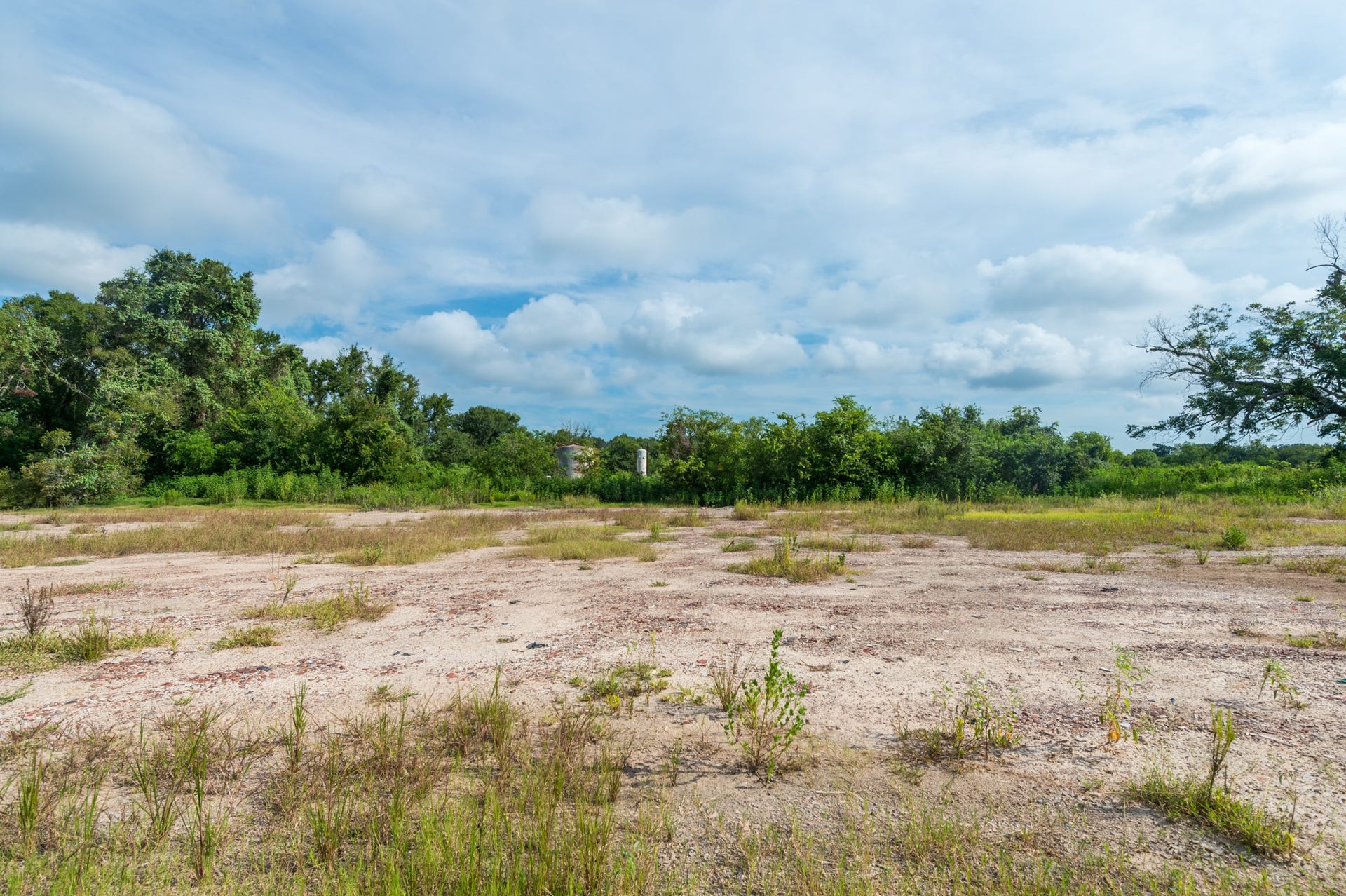 This photo shows a spacious, open plot of land with sparse vegetation and surrounded by lush greenery, under a partly cloudy sky. Ideal for building a home with a natural setting.