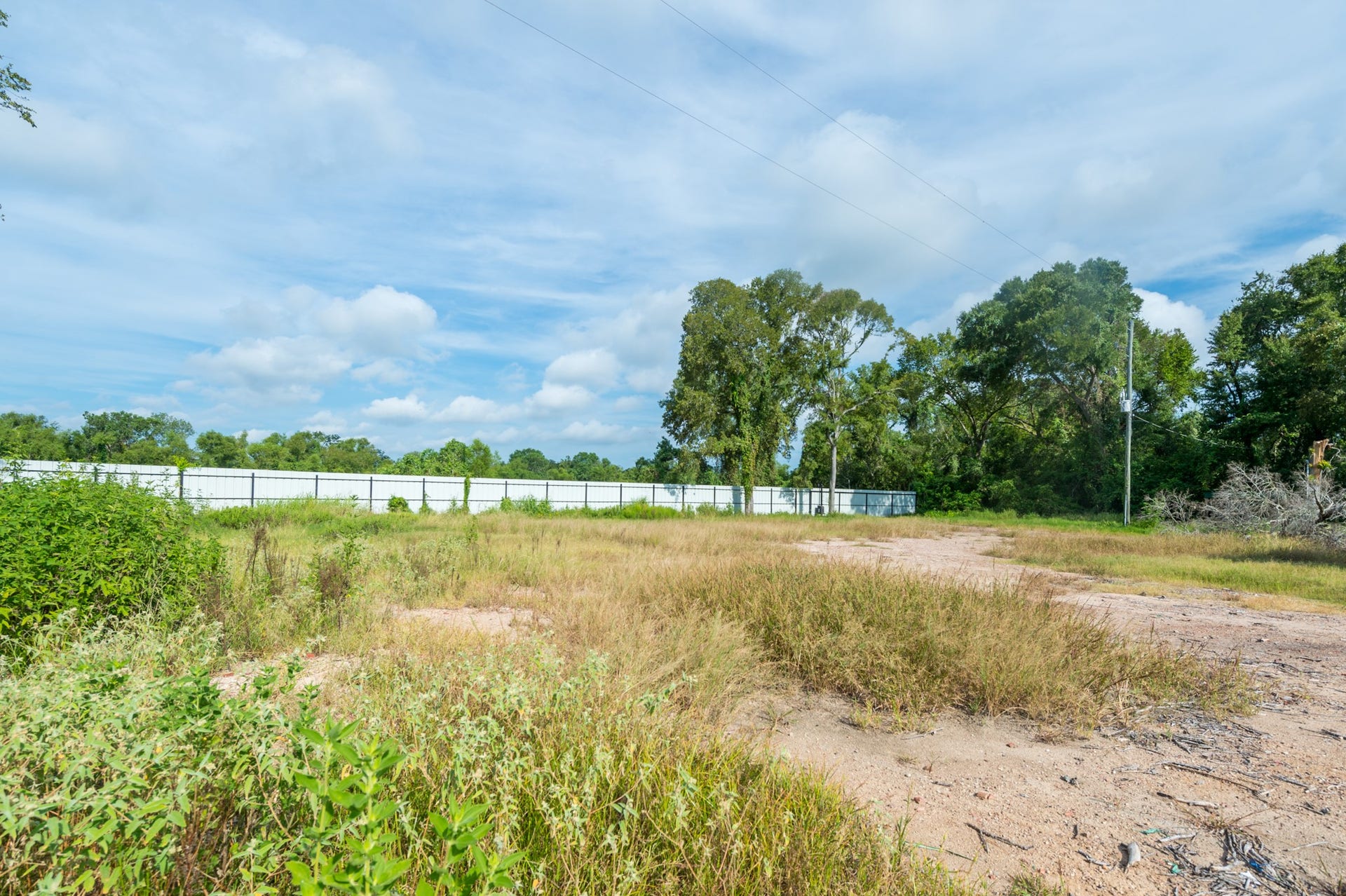 This photo shows a large, open plot of land with some grass and sparse vegetation, bordered by a white fence, and surrounded by trees, offering a spacious and private setting.